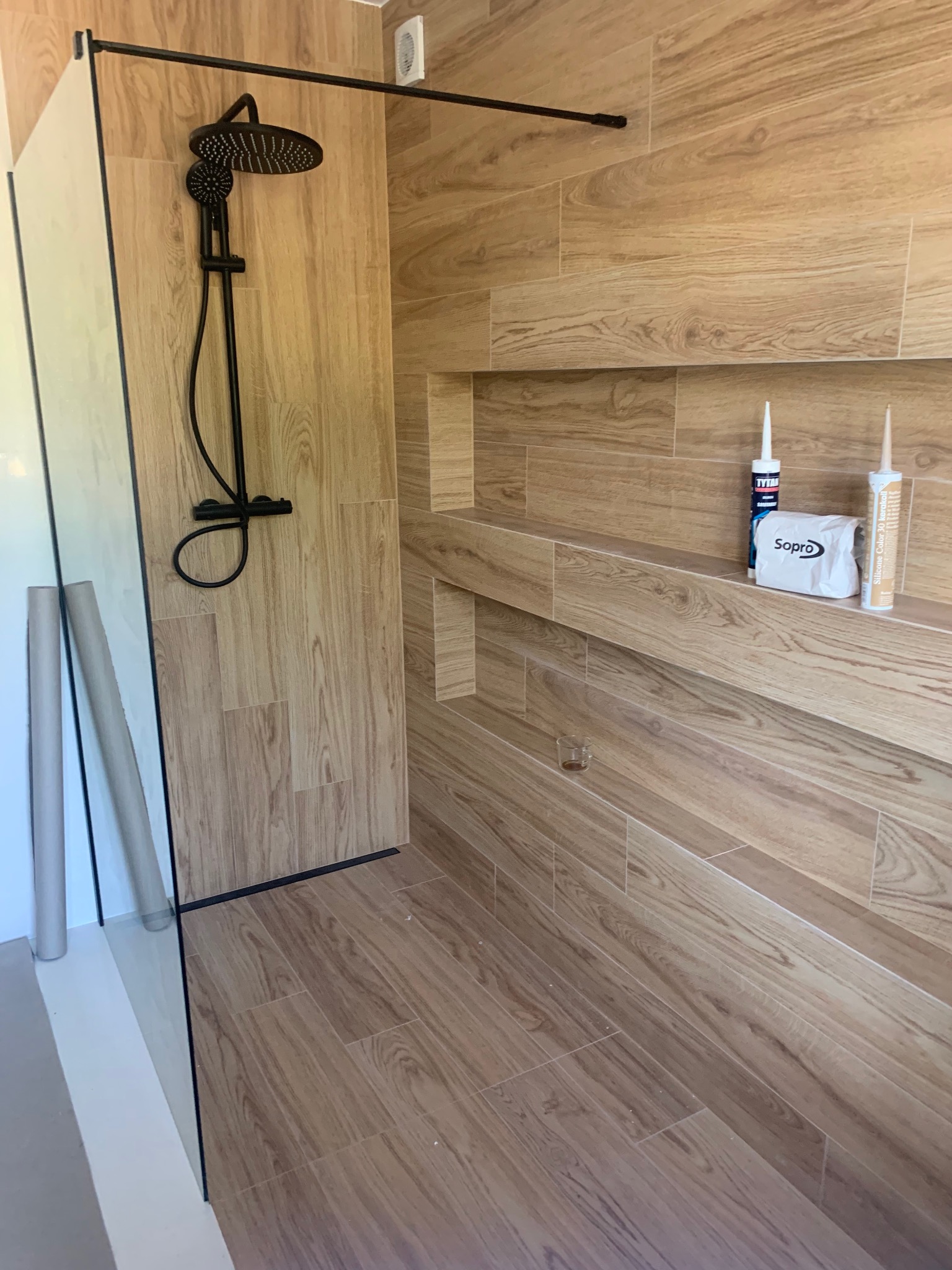 Nowoczesna kabina prysznicowa typu walk-in z czarną armaturą i drewnopodobnymi płytkami, widoczne tuby z silikonem i opakowanie zaprawy Sopro na półce w ścianie.