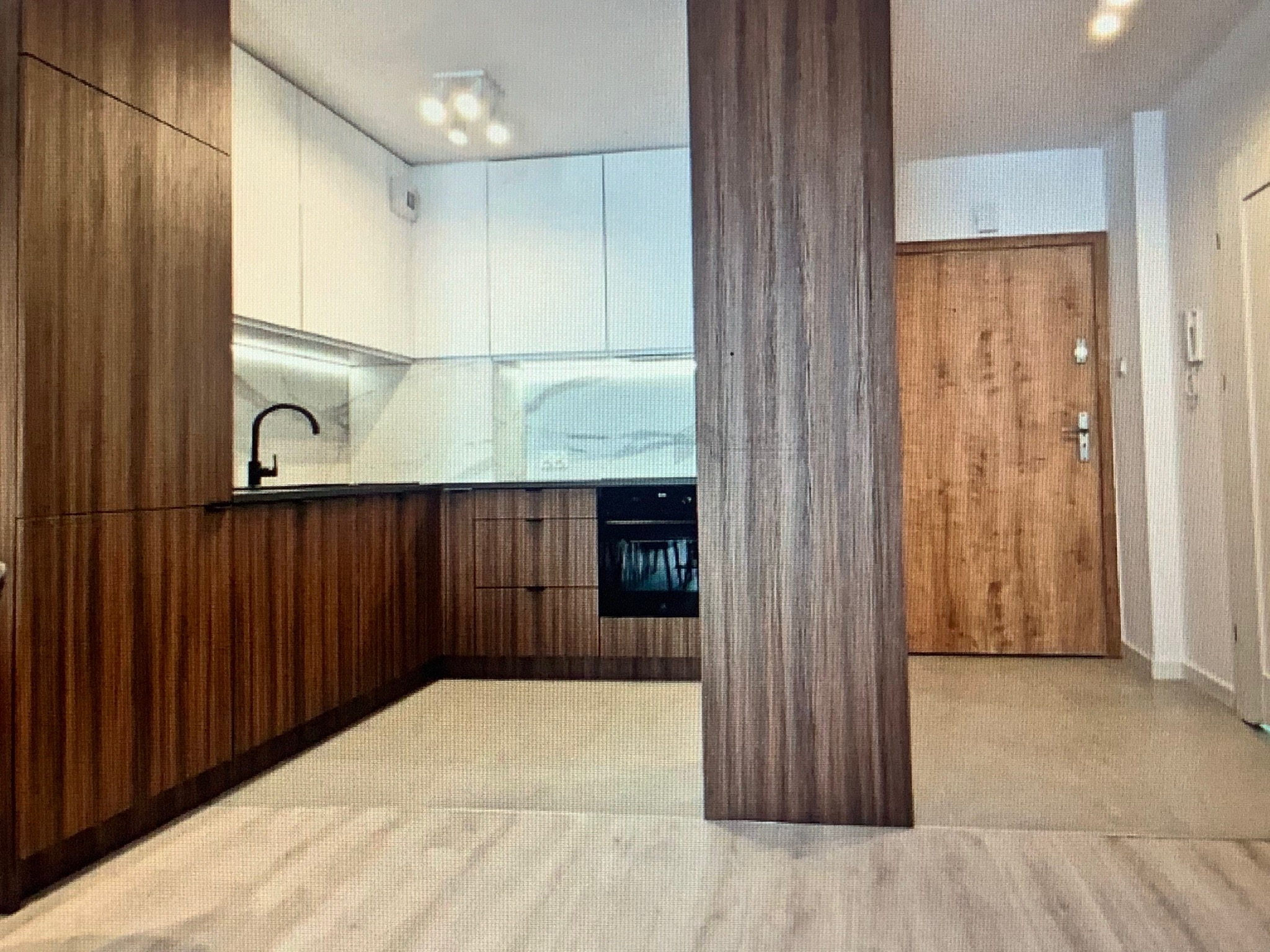 Nowoczesna kuchnia z drewnianymi szafkami, białymi górnymi szafkami i czarną armaturą, widok na drewniane drzwi wejściowe.