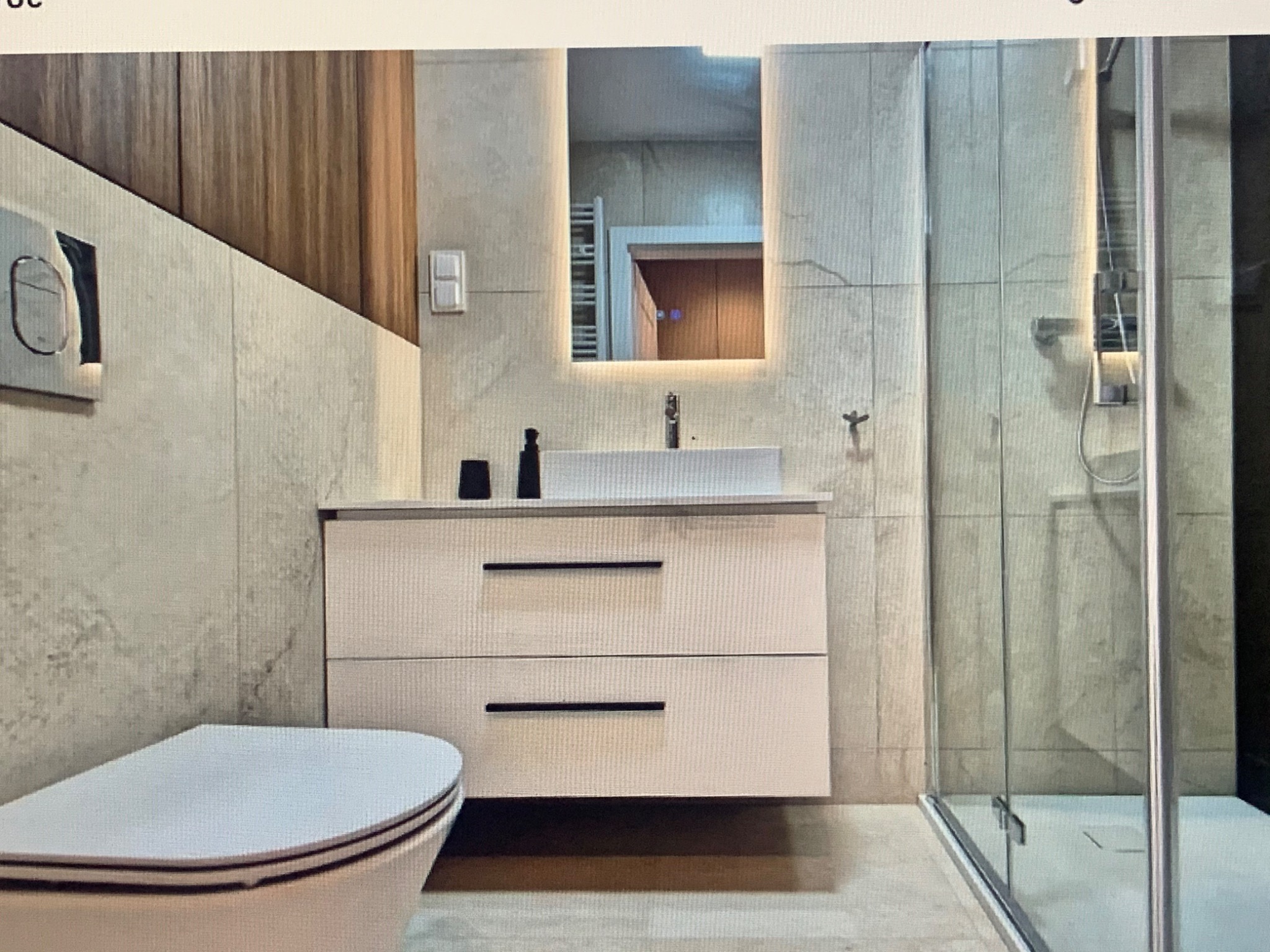 Nowoczesna łazienka z podświetlanym lustrem, białą szafką pod umywalką z czarnymi uchwytami, kabiną prysznicową ze szklanymi drzwiami i toaletą w kolorze białym.