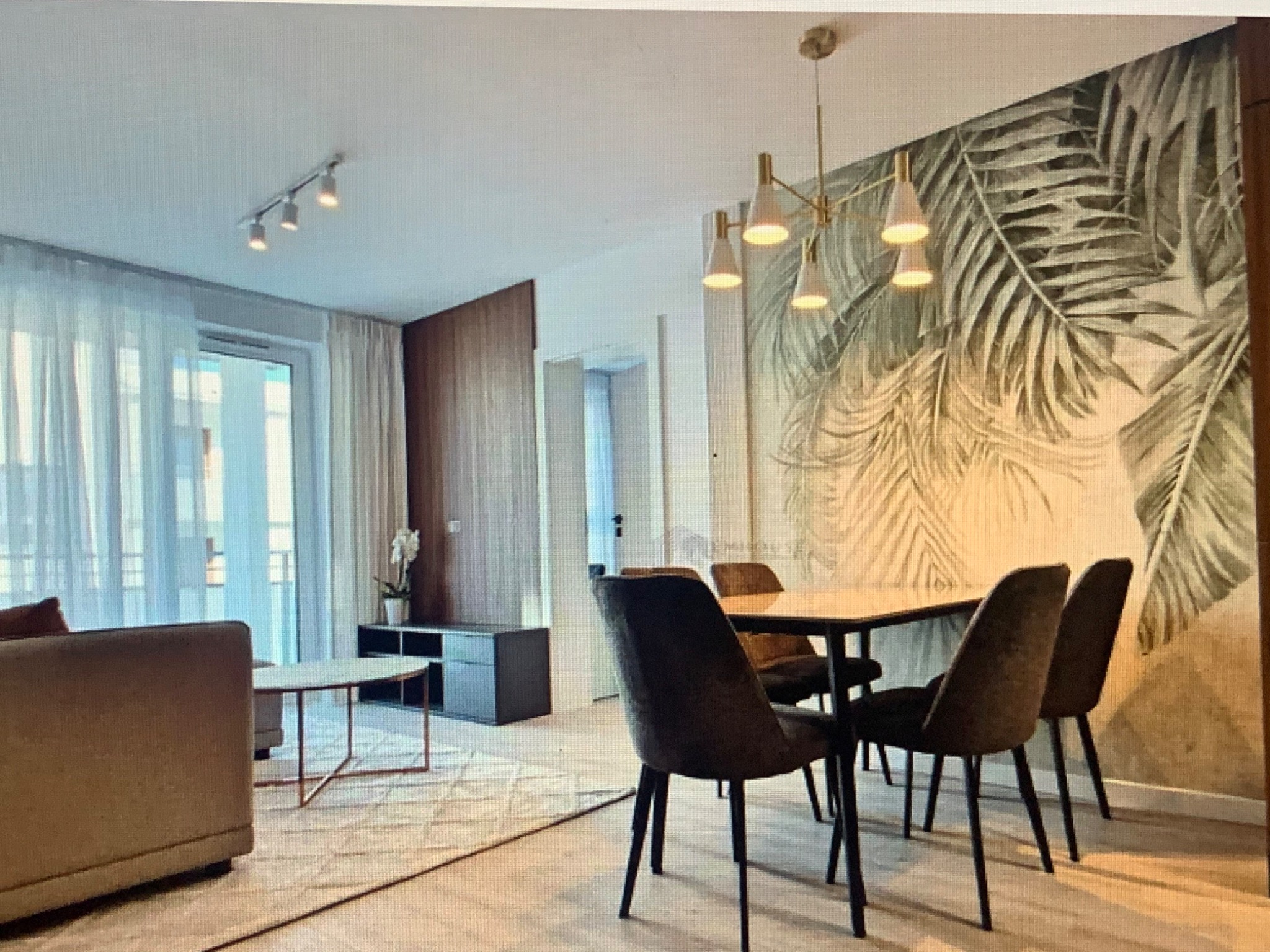 Jasny salon z tapetą w liście palmowe, nowoczesnym oświetleniem i drewnianymi akcentami, widok na balkon przez przesuwne drzwi.