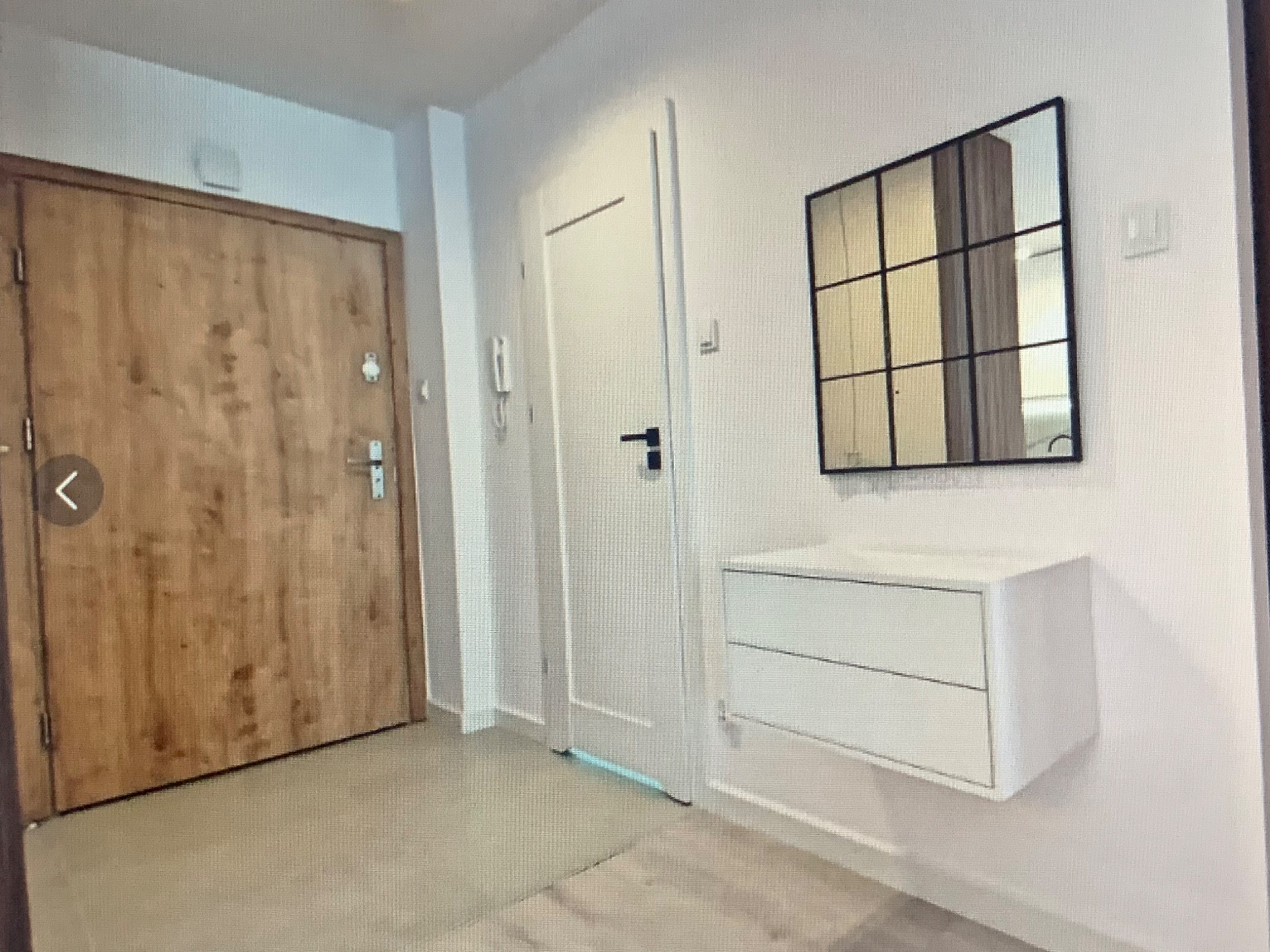 Nowoczesny przedpokój z drewnianymi drzwiami wejściowymi, białymi drzwiami wewnętrznymi, wiszącą szafką z szufladami i lustrem w czarnej ramie z podziałami.