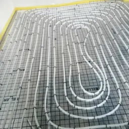 Białe rury ogrzewania podłogowego ułożone w pętle na szarej macie z czarną siatką, zabezpieczone taśmą ż&oacute;łtą przy ścianach, widoczne logo producenta na macie.