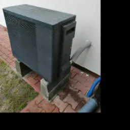 Szara jednostka zewnętrzna, prawdopodobnie klimatyzacji lub pompy ciepła, zamontowana na betonowych bloczkach przy białej ścianie budynku, z widocznymi przewodami odprowadzającymi wodę, na tle...