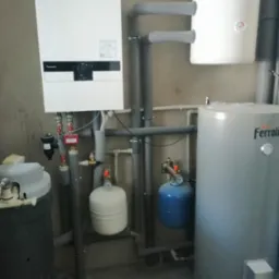 Biały wiszący piec gazowy Panasonic z podłączonymi rurami, obok biały zasobnik ciepłej wody i szary zbiornik Ferroli, instalacja w szarym pomieszczeniu.