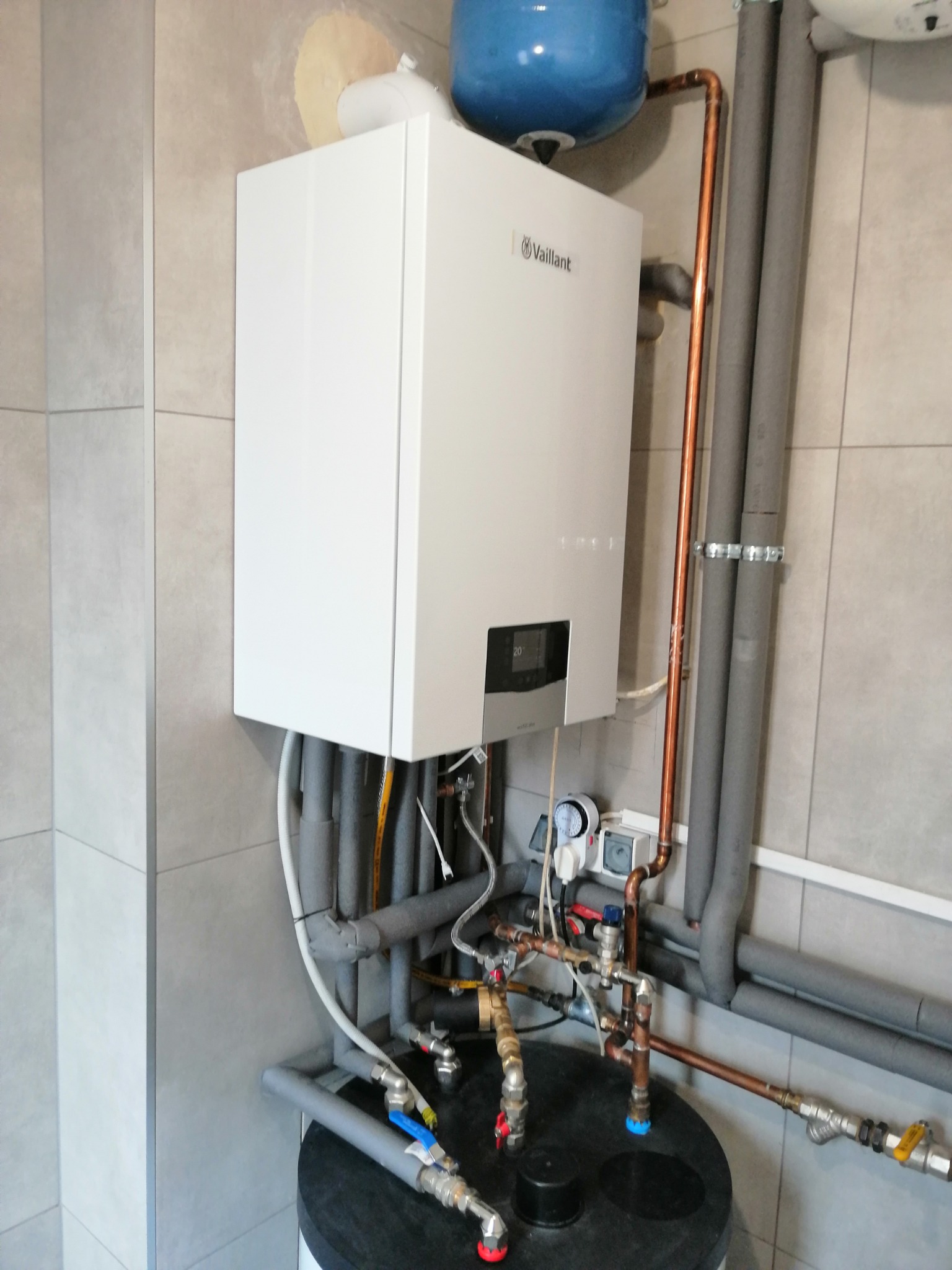 Biały wiszący kocioł gazowy marki Vaillant z podłączonymi rurami miedzianymi i izolowanymi szarymi przewodami, zamontowany na ścianie z szarych płytek, nad czarnym zasobnikiem wody.