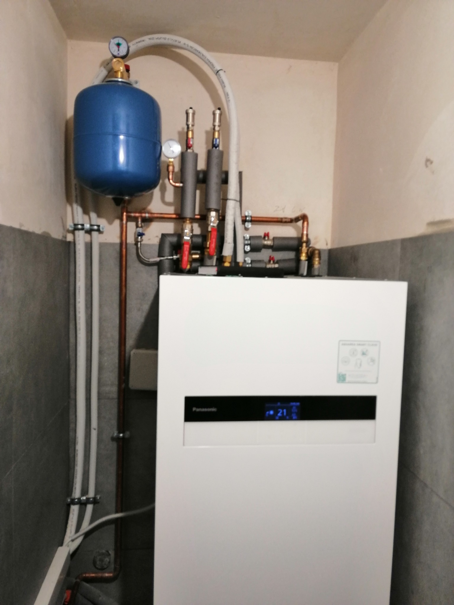 Biała pompa ciepła Panasonic z niebieskim zbiornikiem wyrównawczym i miedzianymi rurami w szarej kotłowni, widoczny wyświetlacz z temperaturą 21 stopni Celsjusza.