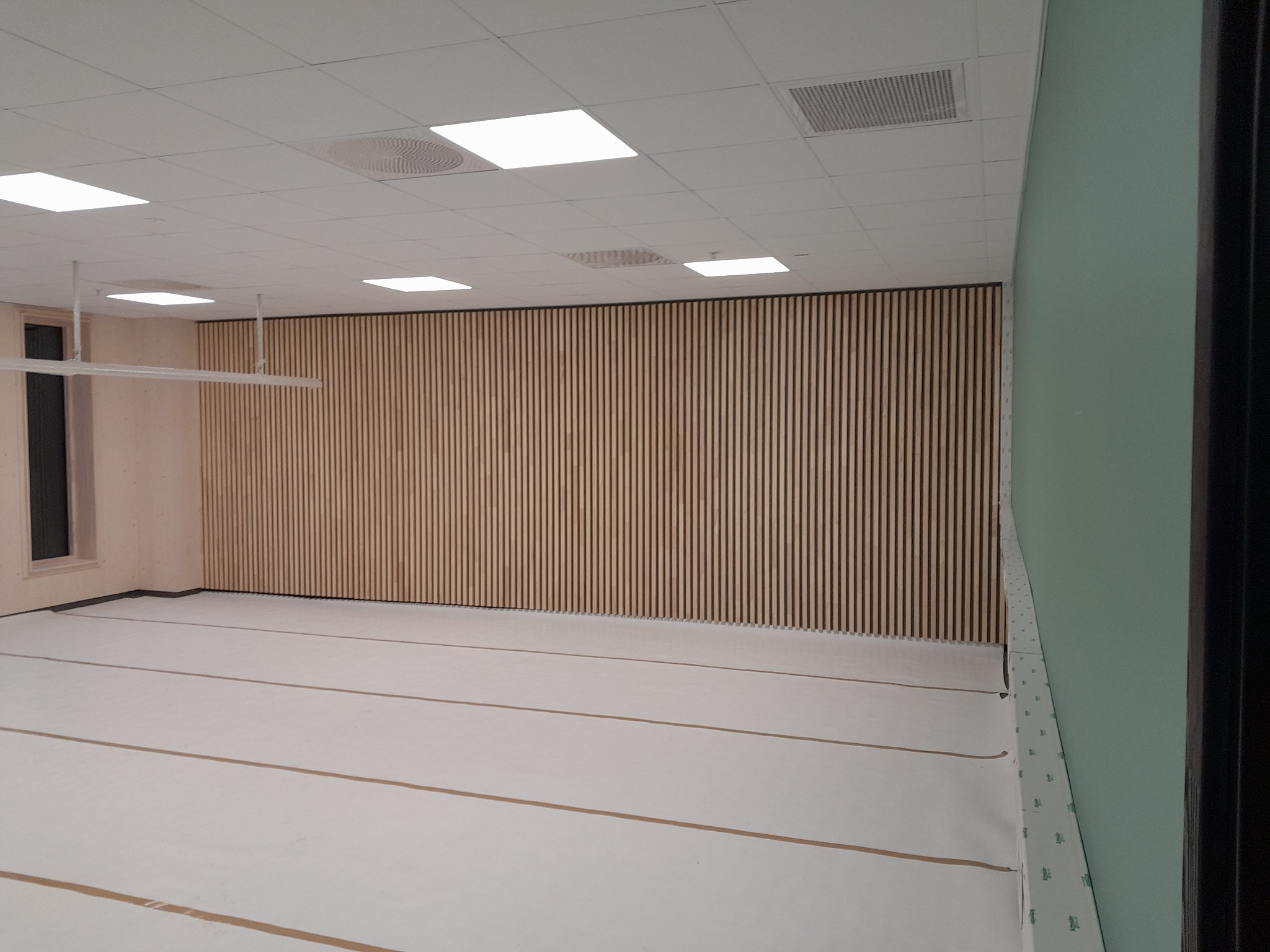 Wnętrze pokoju w trakcie wykańczania: drewniane panele pionowe na ścianie, podłoga przykryta folią ochronną, widoczne elementy instalacji pod sufitem.