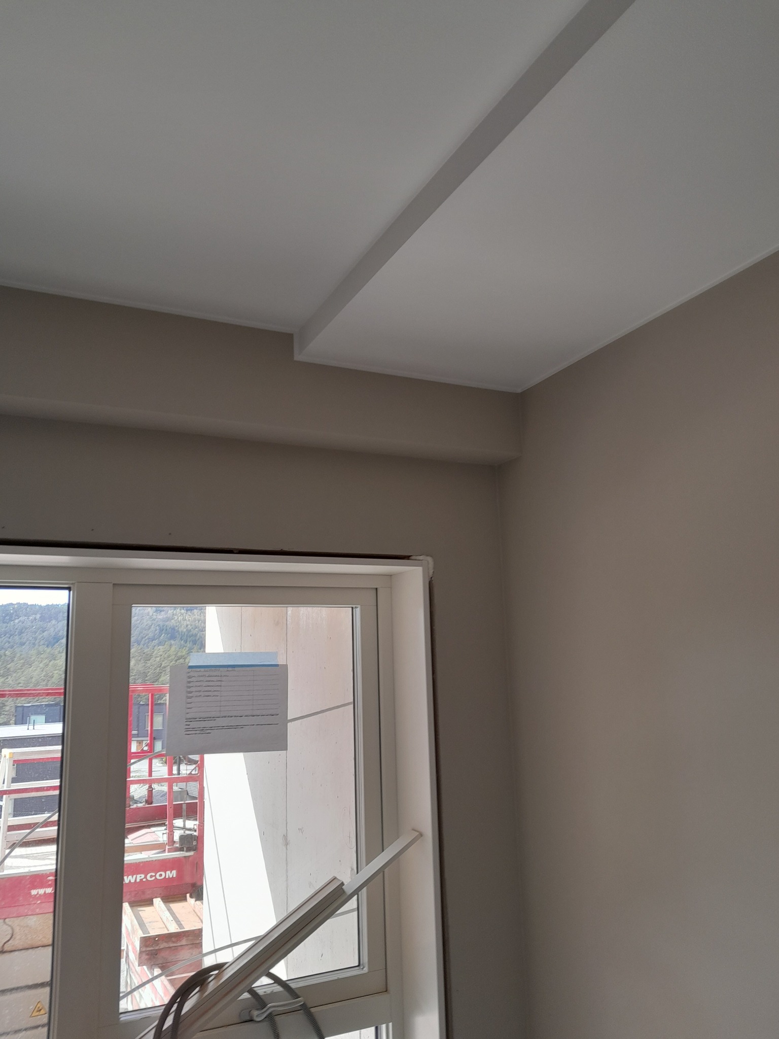 Pomieszczenie po malowaniu: świeżo pomalowane ściany w odcieniach beżu i biały sufit z dekoracyjnym elementem, widok z okna na zewnątrz, gdzie widać podnośnik budowlany.
