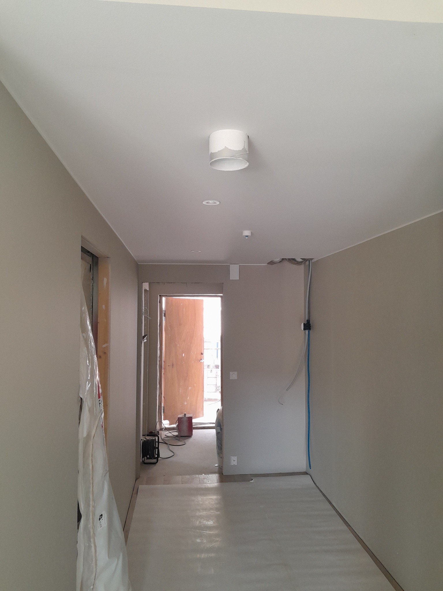 Korytarz w trakcie prac wykończeniowych: zabezpieczona folią ościeżnica, świeżo pomalowane ściany w odcieniach beżu, widoczne przewody elektryczne i biała lampa sufitowa.