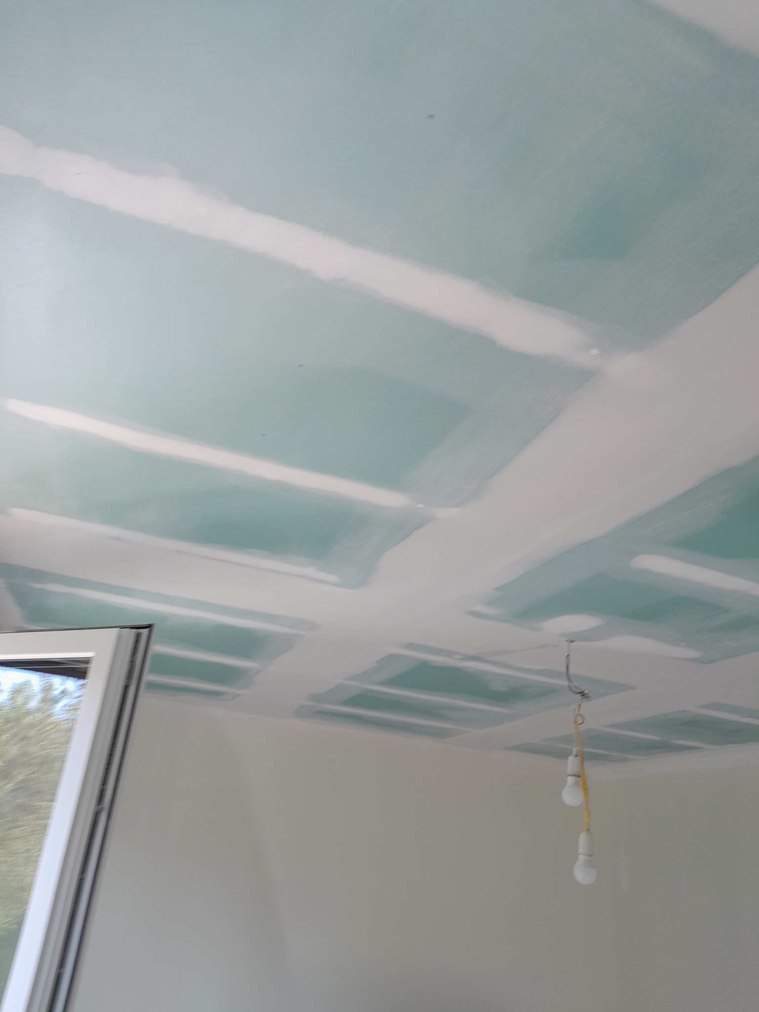 Sufit z płyt gipsowo-kartonowych połączonych masą szpachlową, widoczne niedoskonałości i surowe wykończenie, w tle zwisające żarówki na kablu.