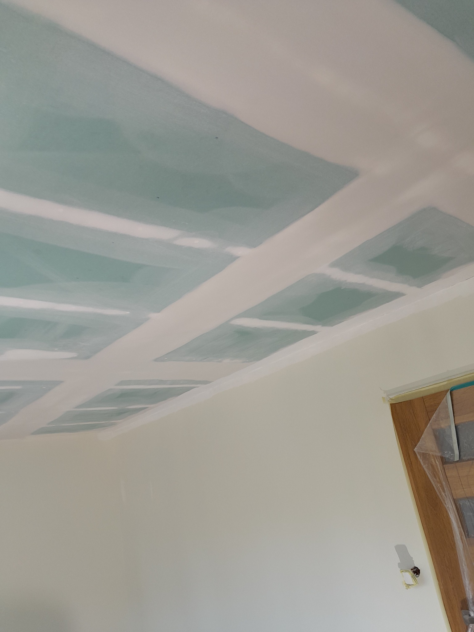 Sufit z płyt gipsowo-kartonowych po szpachlowaniu, widoczne łączenia płyt, przygotowany do dalszych prac wykończeniowych, zabezpieczone drzwi folią malarską.