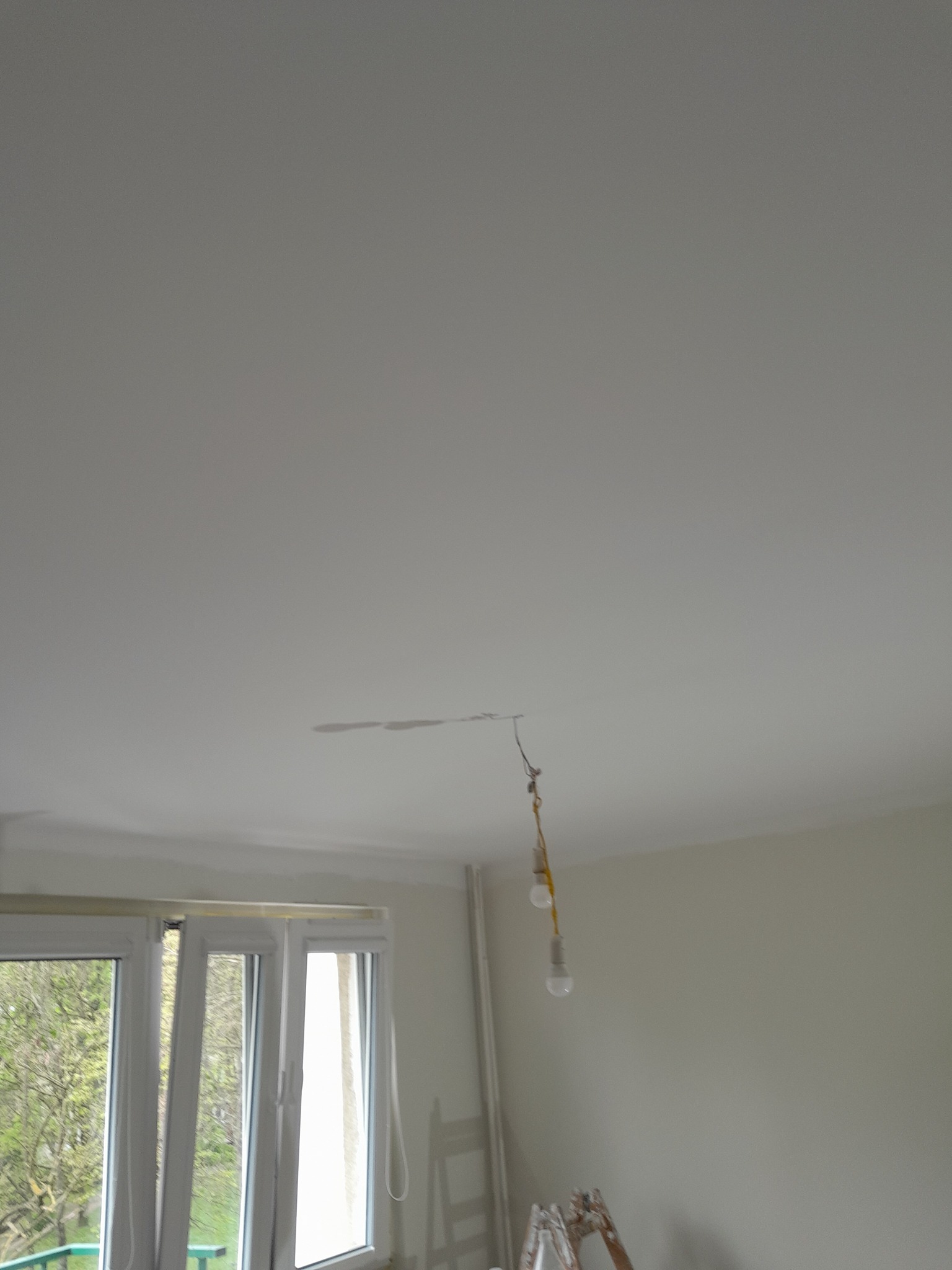 Biały sufit z widocznymi śladami po zalaniu i wiszącymi żarówkami na kablu, w tle okno i drabina malarska.