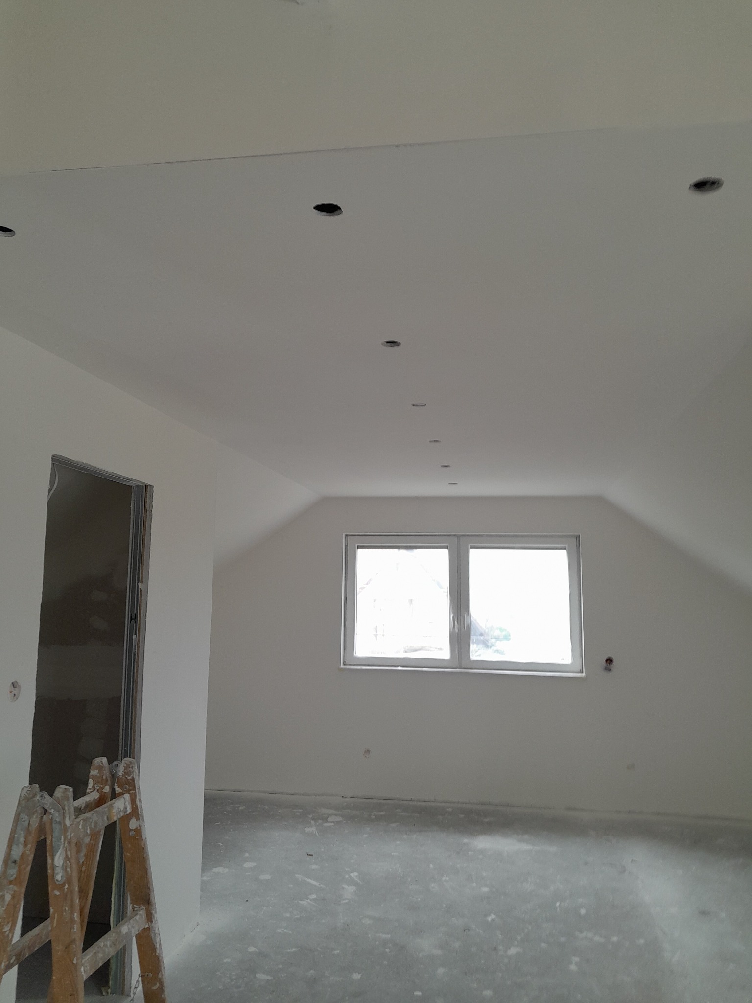 Poddasze w trakcie prac wykończeniowych: białe ściany, sufit z otworami na oświetlenie, okno i drewniana drabina przy otworze drzwiowym, podłoga w stanie surowym.