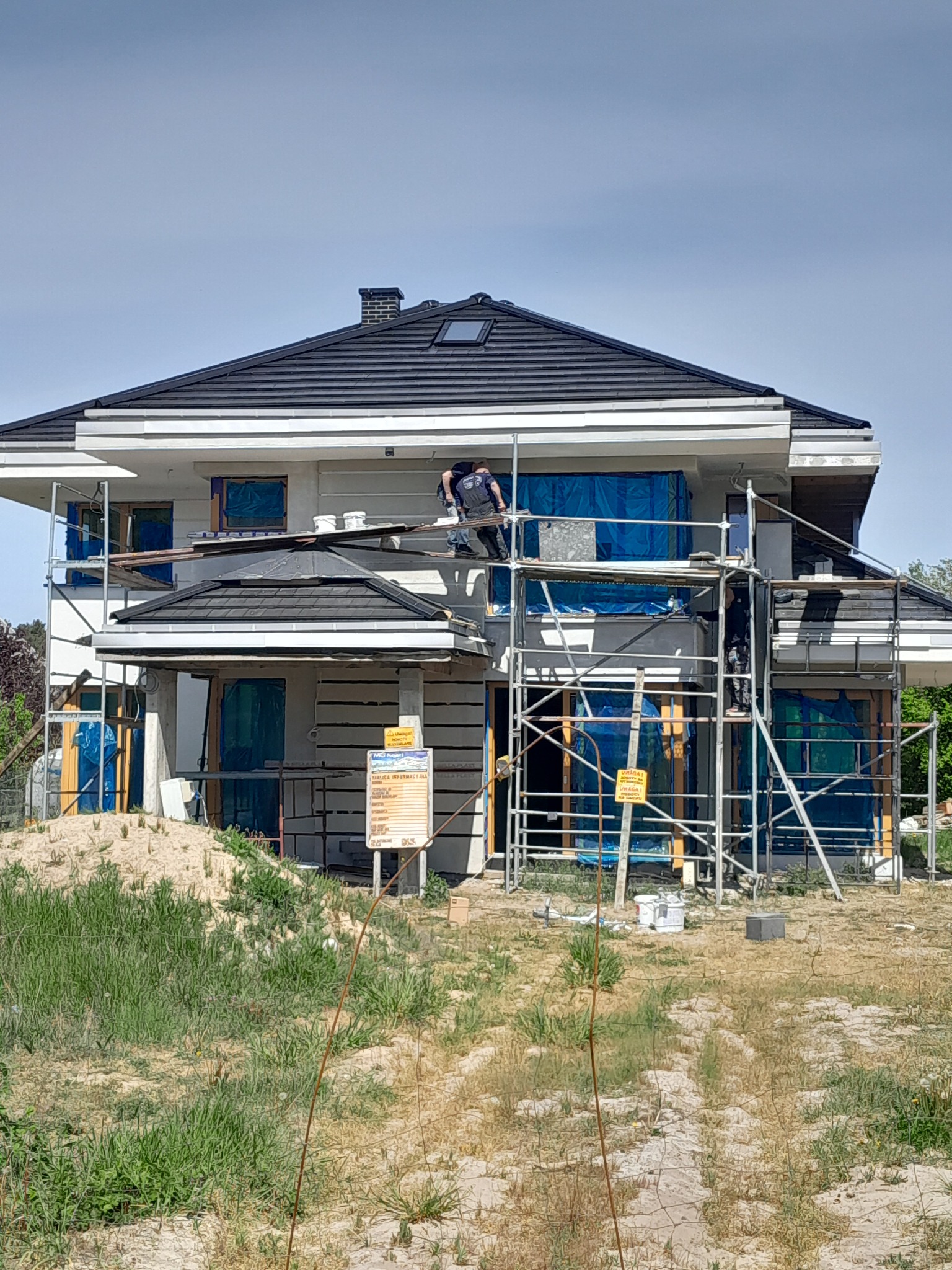 Dom w trakcie prac wykończeniowych w Stargardzie, z rusztowaniami i oknami zabezpieczonymi folią, widoczny pracownik na rusztowaniu oraz przygotowane materiały budowlane.