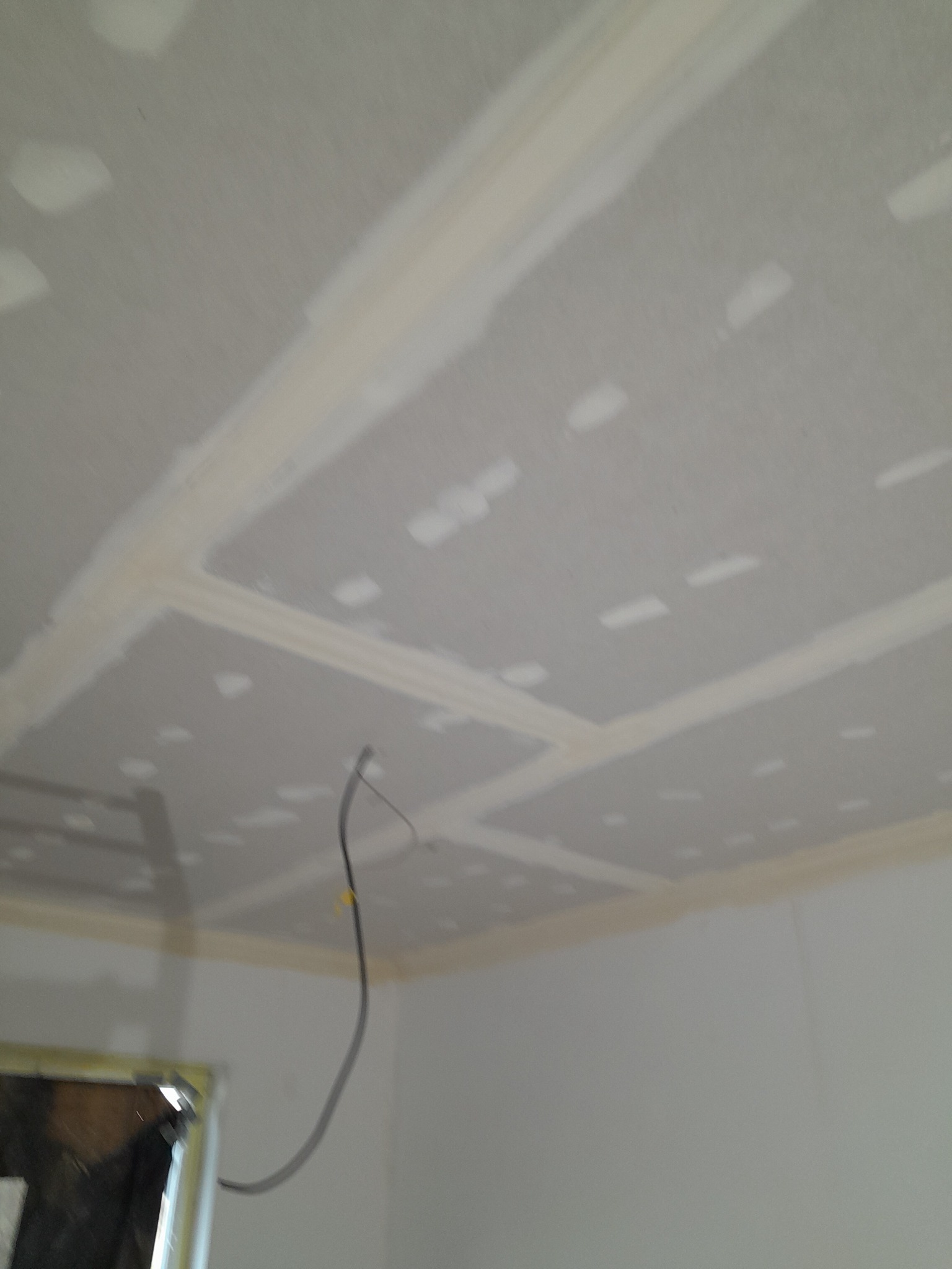 Sufit i fragment ściany po szpachlowaniu płyt gipsowo-kartonowych, widoczne łączenia płyt wypełnione masą szpachlową, kabel elektryczny zwisający z sufitu.