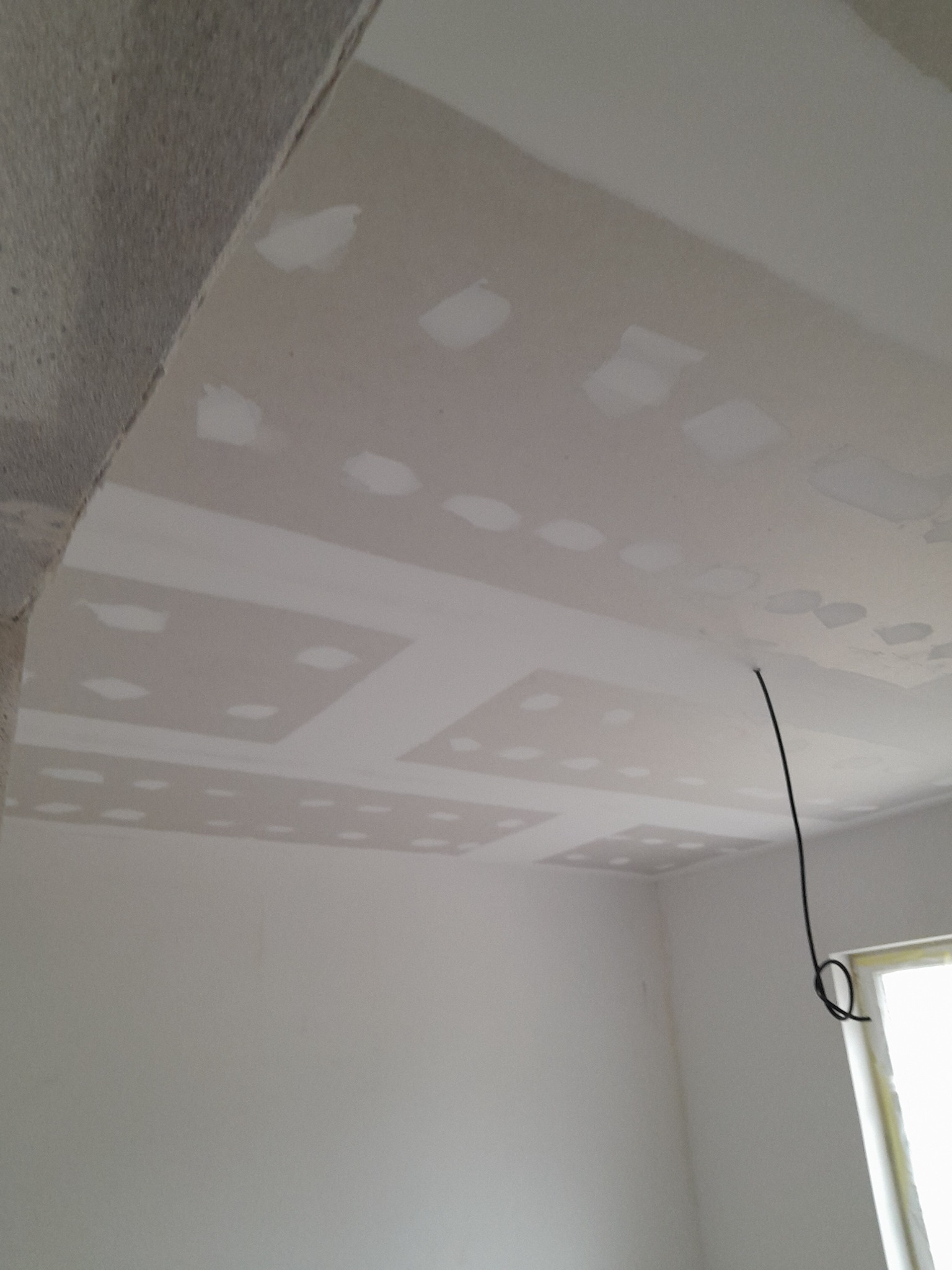 Sufit z płyt gipsowo-kartonowych w trakcie szpachlowania, widoczne miejsca łączeń i wkrętów wypełnione masą szpachlową, surowy betonowy element konstrukcyjny po lewej stronie, instalacja...