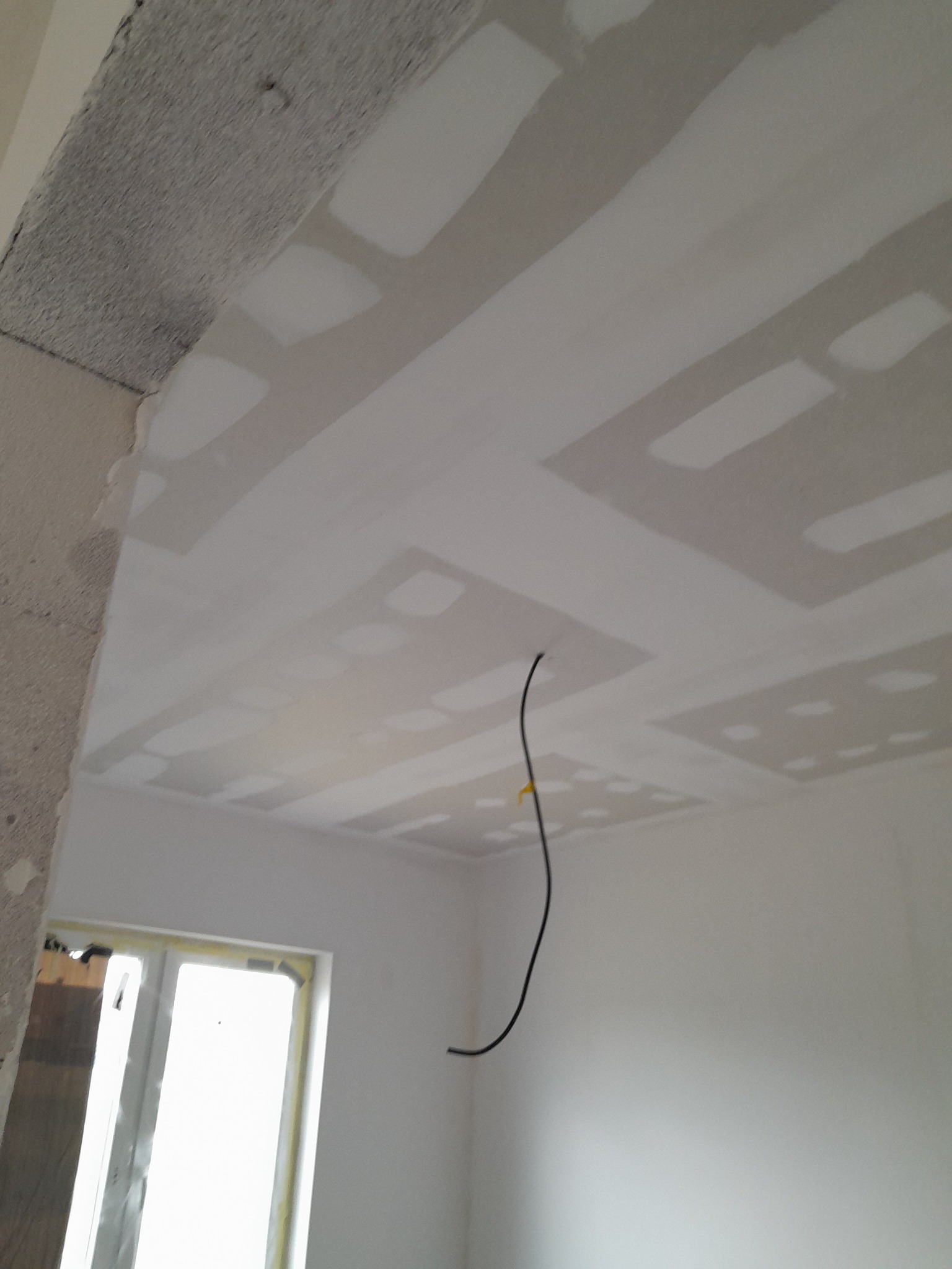 Sufit po szpachlowaniu, z widocznymi łączeniami płyt gipsowo-kartonowych, przygotowany do dalszych prac wykończeniowych, z wiszącym kablem elektrycznym i fragmentem okna w tle.