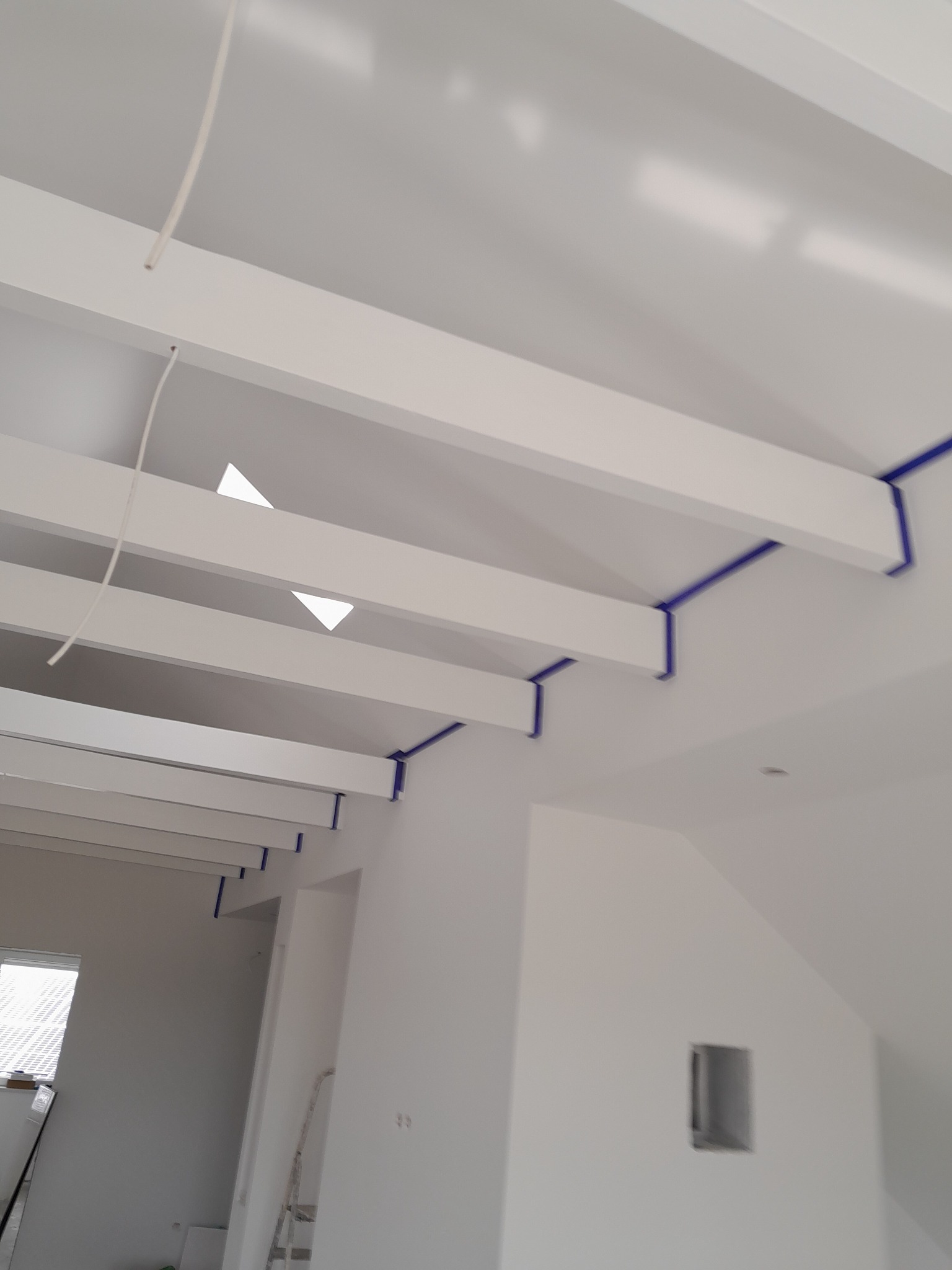 Białe belki stropowe z niebieską taśmą malarską zabezpieczającą, jasne wnętrze w trakcie prac wykończeniowych, widoczna instalacja elektryczna i okno.