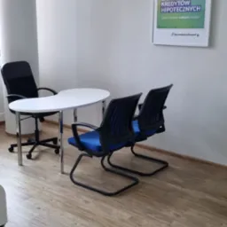 Jasne biuro z okrągłym białym stołem, trzema krzesłami (dwa z niebieskimi siedziskami), kolumną i plakatem na ścianie 'Sprawdzony Ekspert.pl - Specjaliści do spraw kredyt&oacute;w hipotecznych'.