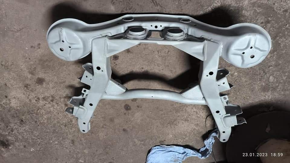 Element podwozia samochodowego po renowacji, pokryty jasnoszarą farbą proszkową, widoczne otwory montażowe, na szarym betonowym podłożu z widocznym fragmentem niebieskiej szmatki.