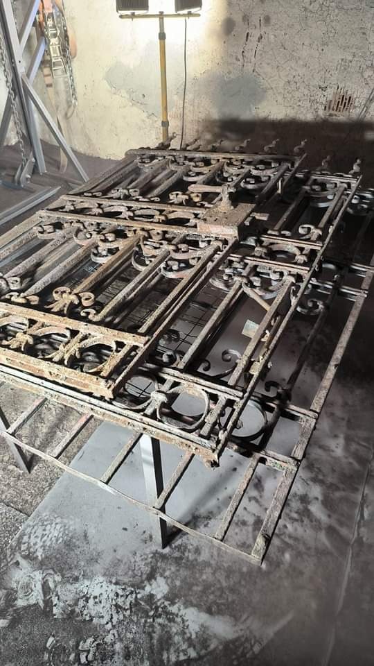 Metalowe elementy balustrady z dekoracyjnymi zdobieniami, pokryte rdzą, ułożone na metalowym stelażu. W tle widoczna lampa warsztatowa i ściana z odpryskującą farbą.