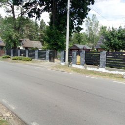 Gol -Bud - Nowoczesne, poziome ogrodzenie z czarnych desek i szarych betonowych słupków, widok z ulicy, z żółtą skrzynką gazową przy jednym ze słupków.