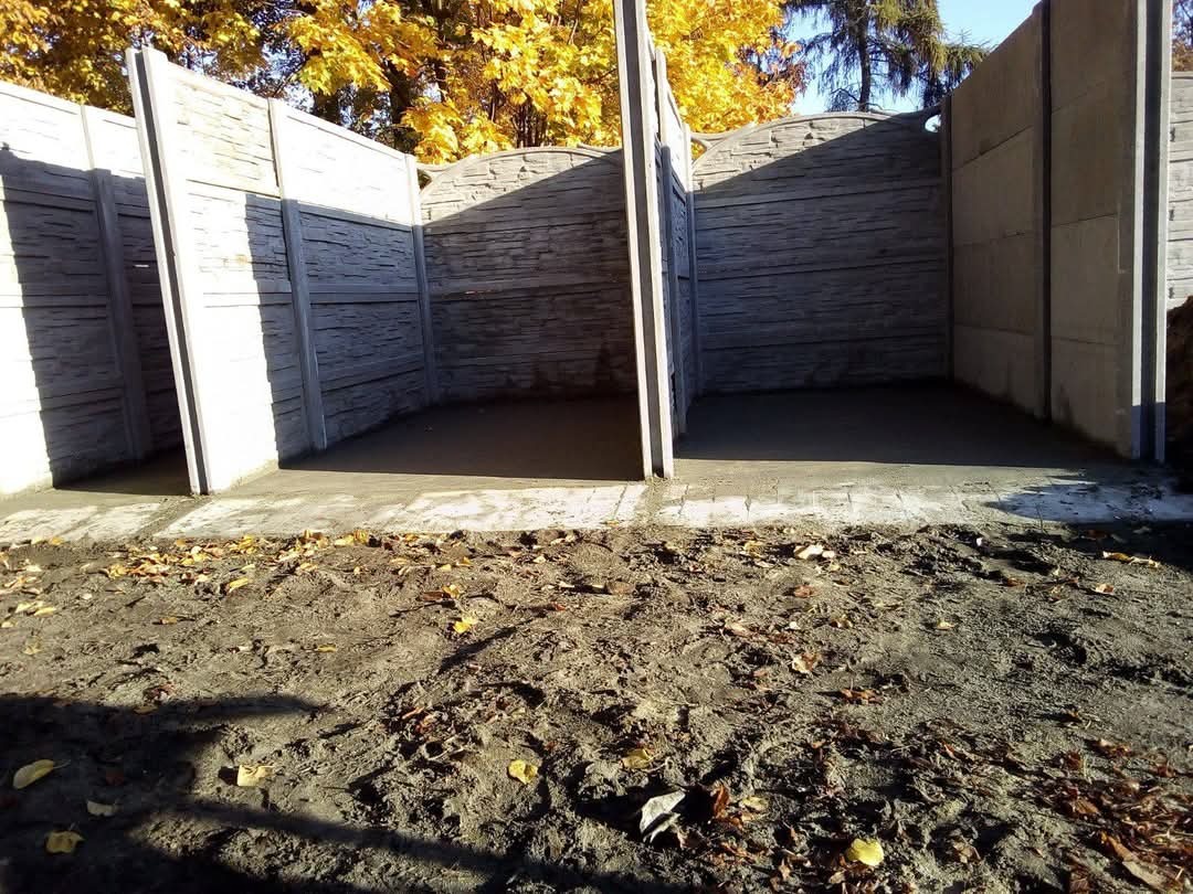 Fragment betonowego ogrodzenia panelowego z widocznymi słupkami i wypełnieniami, teren przygotowany pod wylewkę, jesienne liście na ziemi.