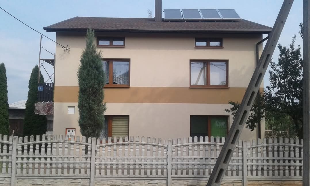 Dwukondygnacyjny dom z panelami solarnymi na dachu, ogrodzony betonowym płotem z ozdobnymi łukami i filarami, widoczny słup elektryczny oraz rusztowanie z boku budynku.