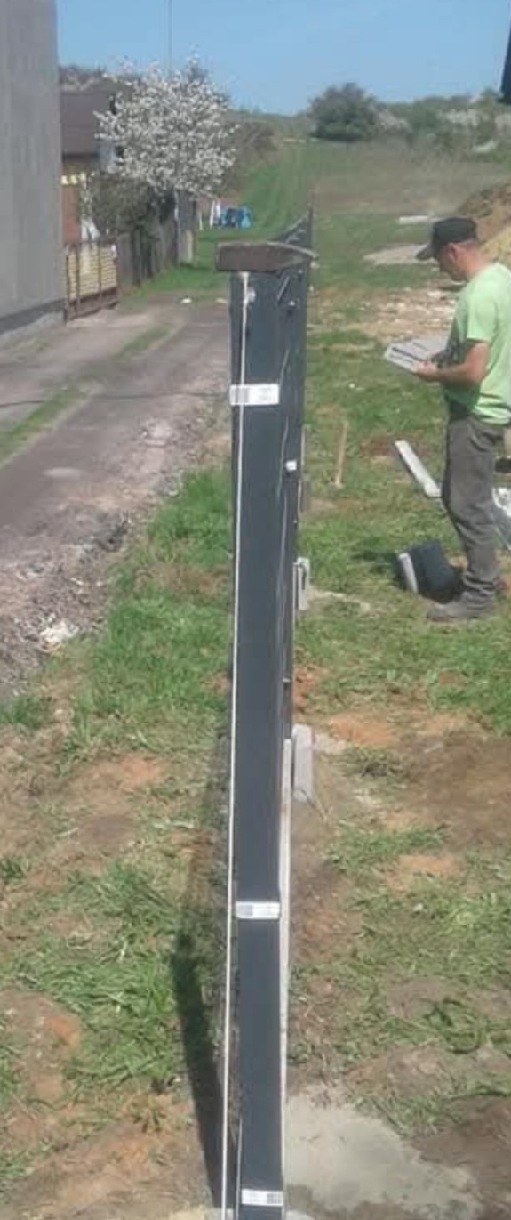 Wkopany w ziemię słupek ogrodzeniowy z przymocowanymi elementami montażowymi, w tle majster w zielonej koszulce podczas pracy.