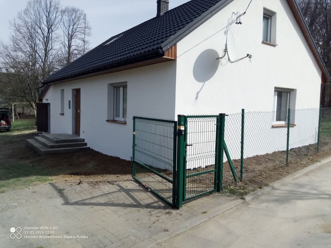 Otwarta, zielona furtka z siatki panelowej na tle białego domu z czarnym dachem i zielonego ogrodzenia z siatki plecionej, widok z zewnątrz.