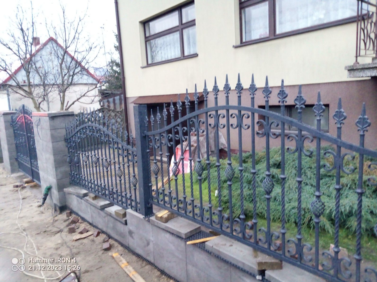 Fragment montowanego, ozdobnego, metalowego ogrodzenia z elementami kutymi, widoczne słupki z szarej kostki i podmurówka, w tle budynek mieszkalny.