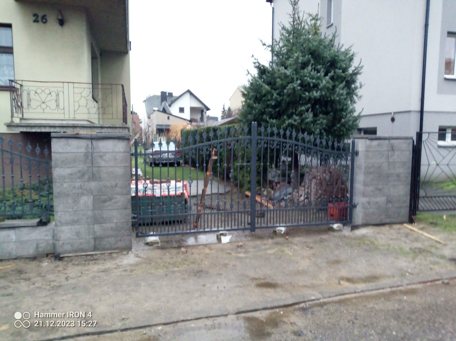 Nowoczesna, czarna brama ogrodzeniowa z dekoracyjnymi elementami, osadzona na szarych, betonowych słupkach, widoczna na tle domu z numerem 26 i zielonego świerku, w trakcie montażu; na ziemi...