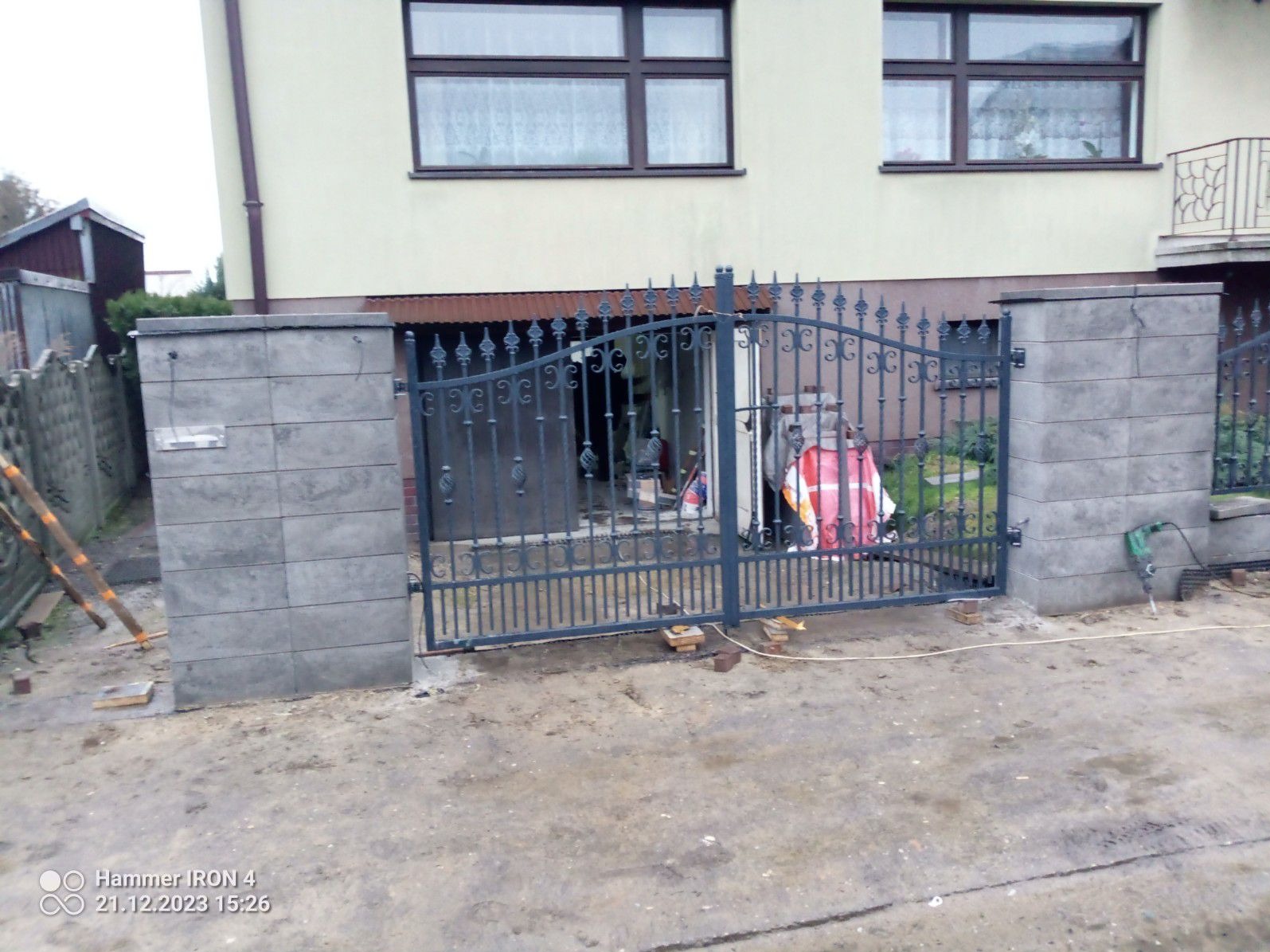 Nowo zamontowana, metalowa brama wjazdowa z ozdobnymi elementami, osadzona między dwoma murowanymi słupkami z szarej kostki, widoczny budynek w tle i narzędzia pracy.