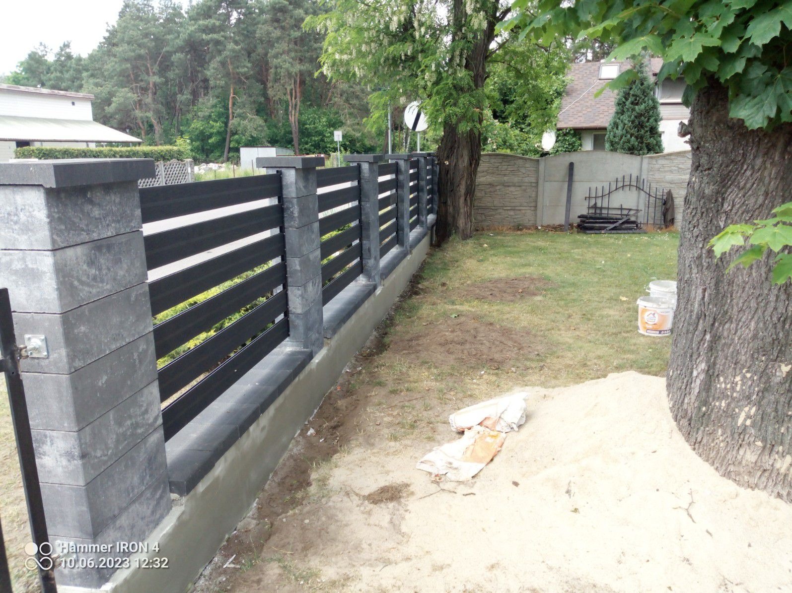 Nowoczesne ogrodzenie z poziomych, czarnych paneli i szarych, betonowych słupków, z widocznymi śladami świeżego betonu u podstawy, na tle trawnika i drzew.