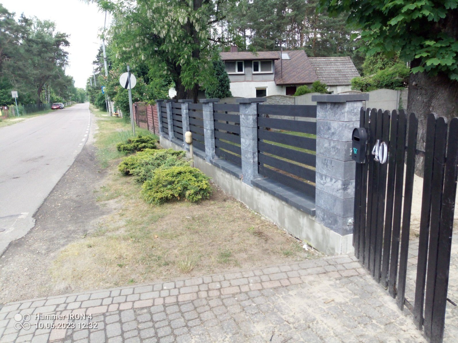 Nowoczesne ogrodzenie panelowe z betonowymi słupkami i czarną furtką z numerem 40, widok z chodnika na tle ulicy i domu.