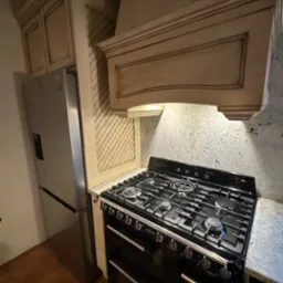 Kuchnia w stylu rustykalnym: zabudowa kuchenna z kremowymi szafkami, okapem i wzorzystym panelem ściennym obok srebrnej lodówki SMEG, z czarną, dużą kuchenką gazową.