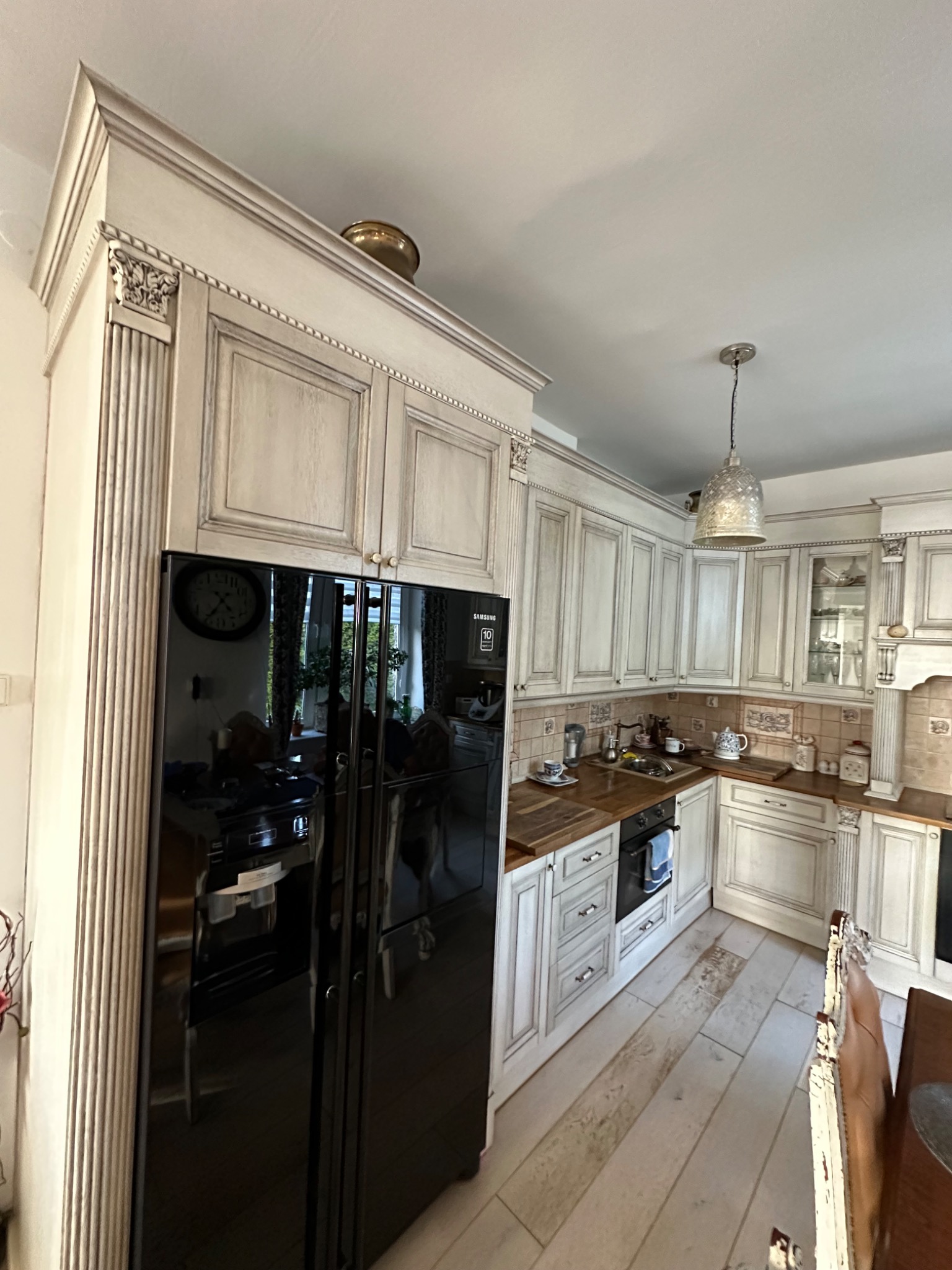 Rustykalna kuchnia z kremowymi frontami szafek, drewnianymi blatami i czarną lodówką Samsung z widocznym odbiciem wnętrza, oświetlona wiszącą lampą.