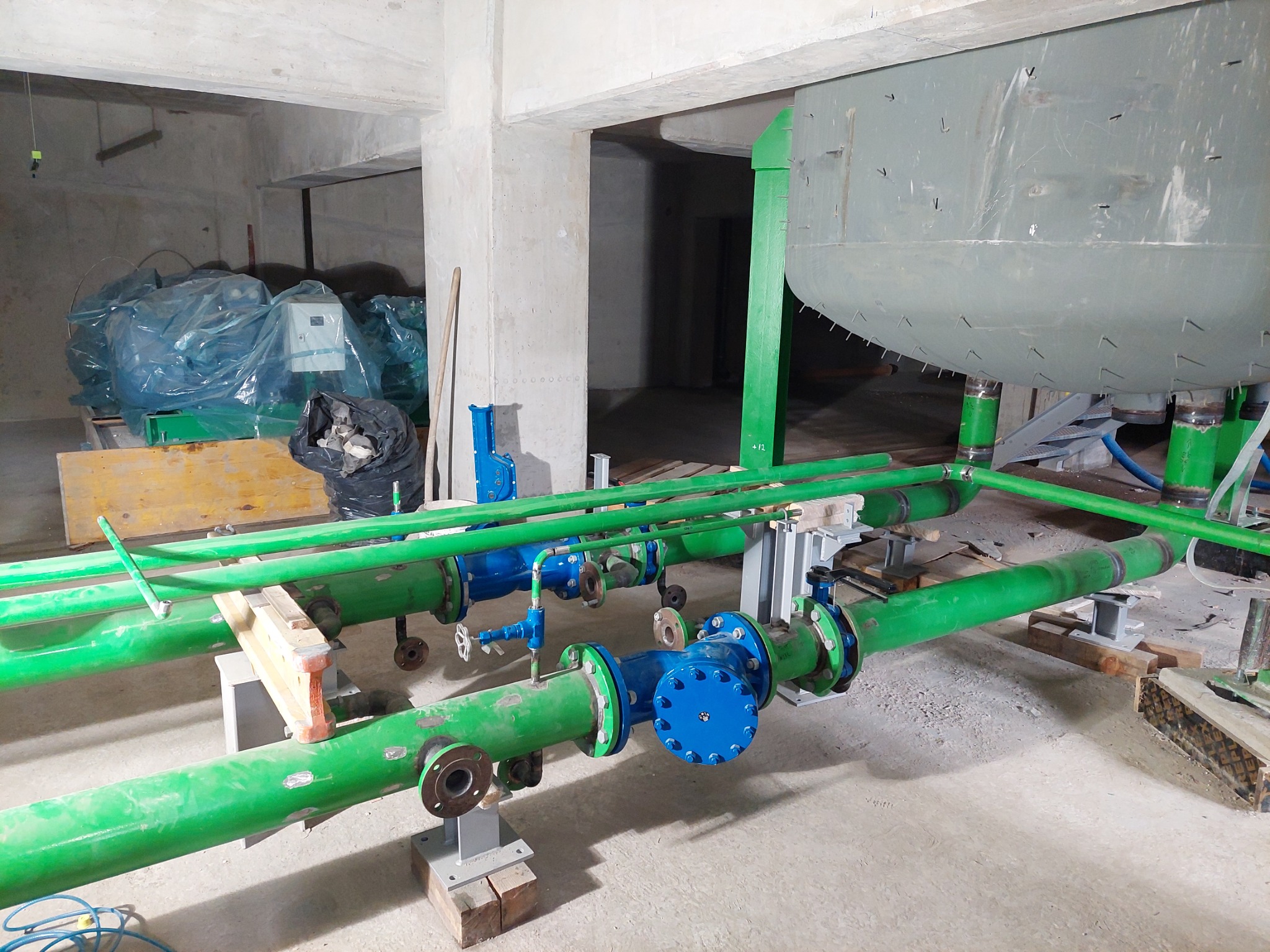 Wnętrze hali w surowym stanie z widoczną konstrukcją zbiornika i systemem rurociągów w kolorze zielonym i niebieskim, wspartych na drewnianych i metalowych podporach. Na podłodze widoczne ślady...