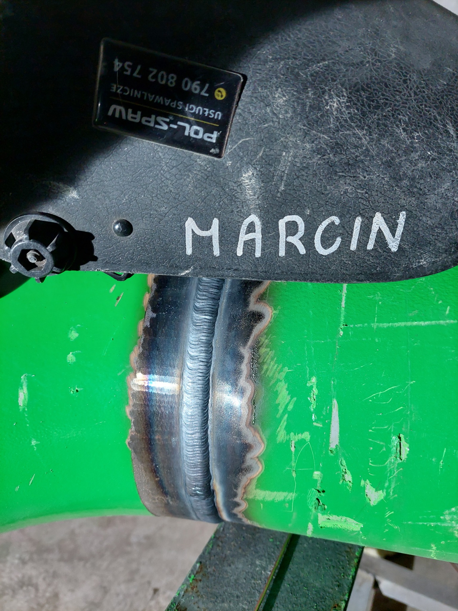 Spoina na zielonej rurze, widoczna spod maski spawalniczej z napisem 'MARCIN' i naklejką 'POL-SPAW USŁUGI SPAWALNICZE 790 802 754'.