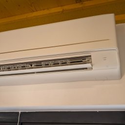 Biały klimatyzator Panasonic zamontowany na ścianie w pomieszczeniu z drewnianym sufitem. Widoczne detale urządzenia i otoczenia.