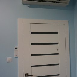 Biały klimatyzator Panasonic zamontowany nad nowoczesnymi, białymi drzwiami z poziomymi panelami na błękitnej ścianie. Widoczny pilot i włącznik światła.