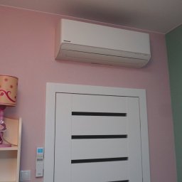 Biały klimatyzator Panasonic na różowej ścianie pokoju dziecięcego, nad białą ramą drzwi. Widoczny pilot i lampka nocna z motywem księżniczki.