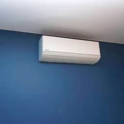 artermo.pl Arkadiusz Klukowski - Biały klimatyzator Panasonic zamontowany na niebieskiej ścianie w pokoju z nowoczesnym oświetleniem. Widok z dołu, podkreślający estetykę instalacji.