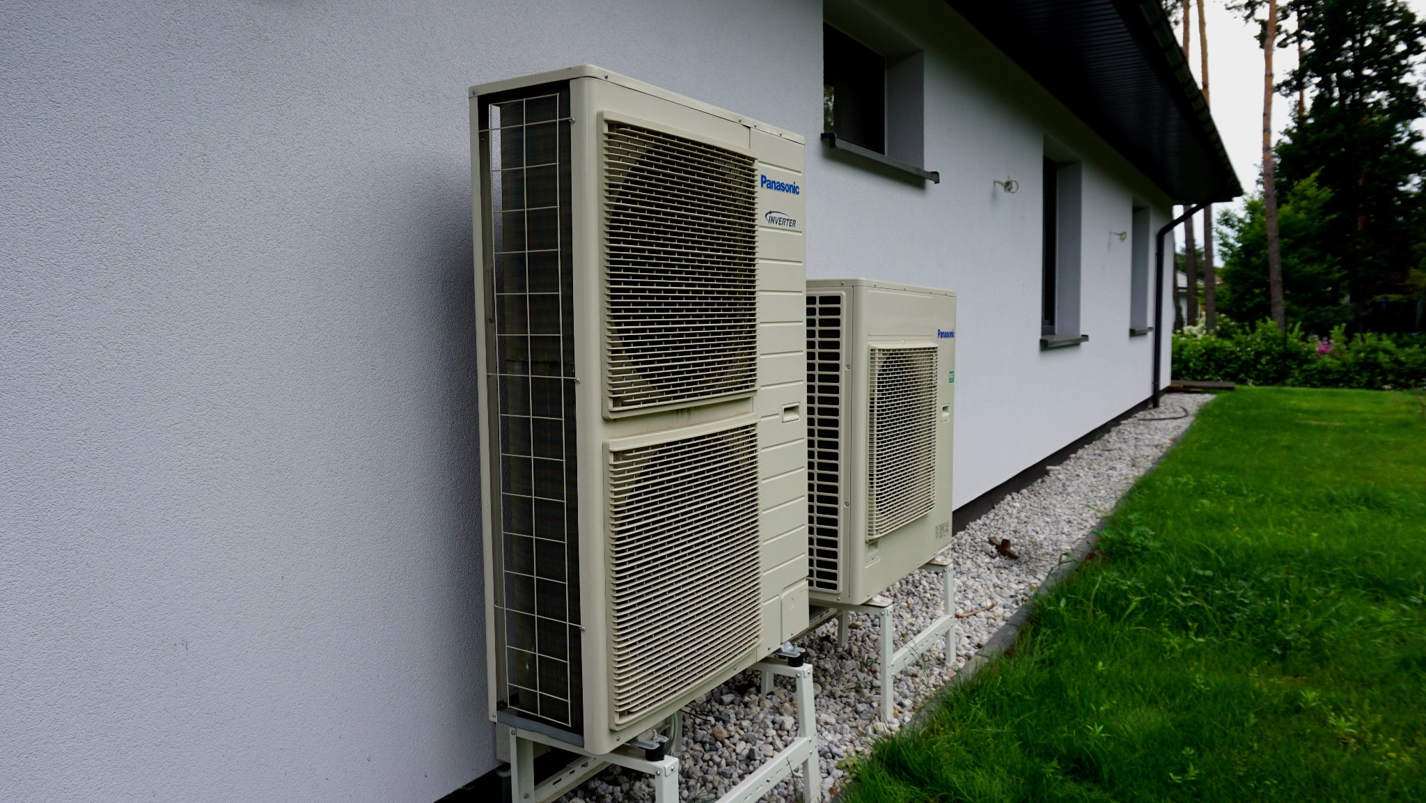 Dwa klimatyzatory Panasonic Inverter zamontowane na zewnątrz białego budynku z zielonym trawnikiem. Widoczne okna i drzewa w tle. Ujęcie z boku, z bliska.
