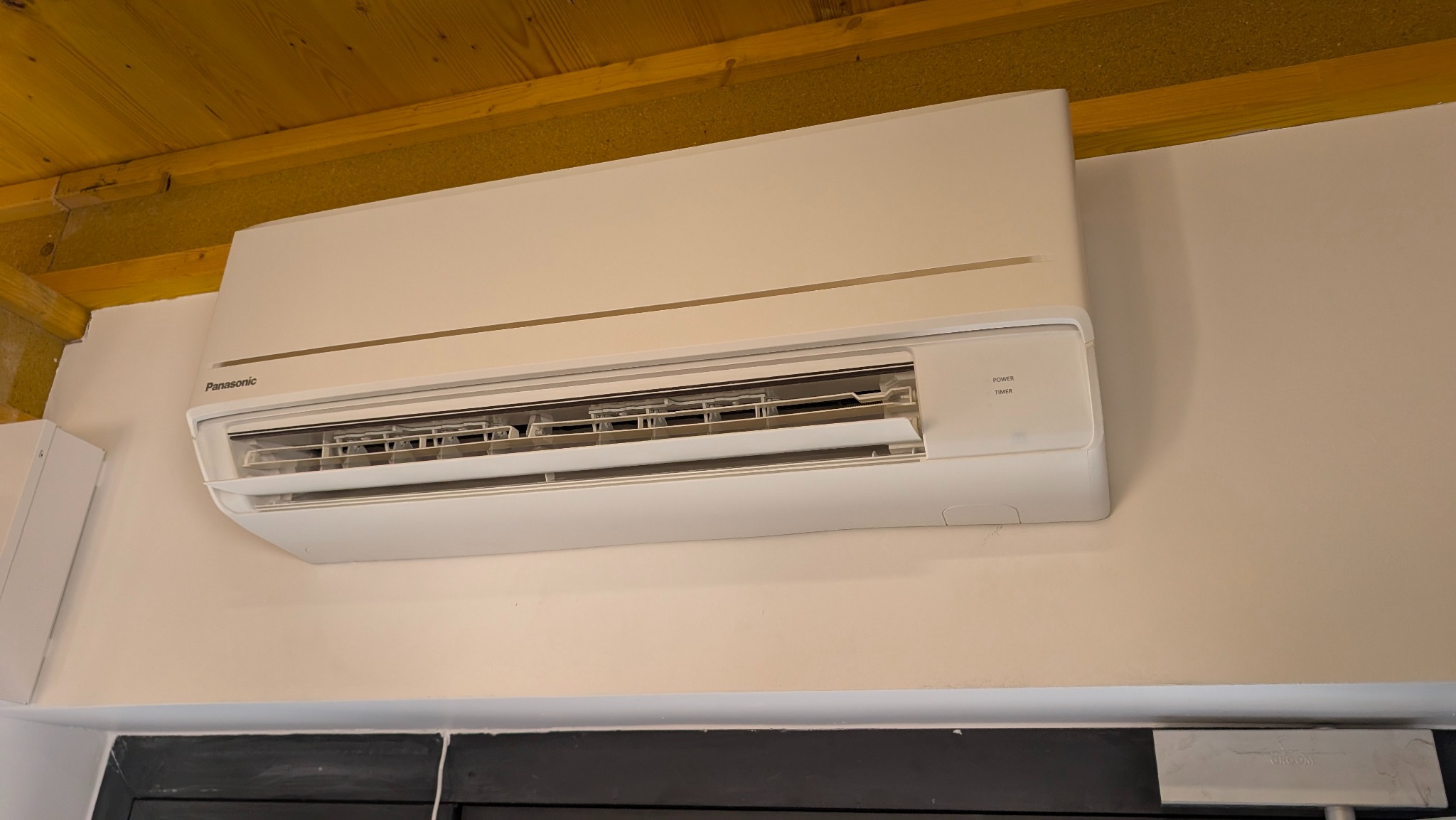 Biały klimatyzator Panasonic zamontowany na ścianie w pomieszczeniu z drewnianym sufitem. Widoczne detale urządzenia i otoczenia.