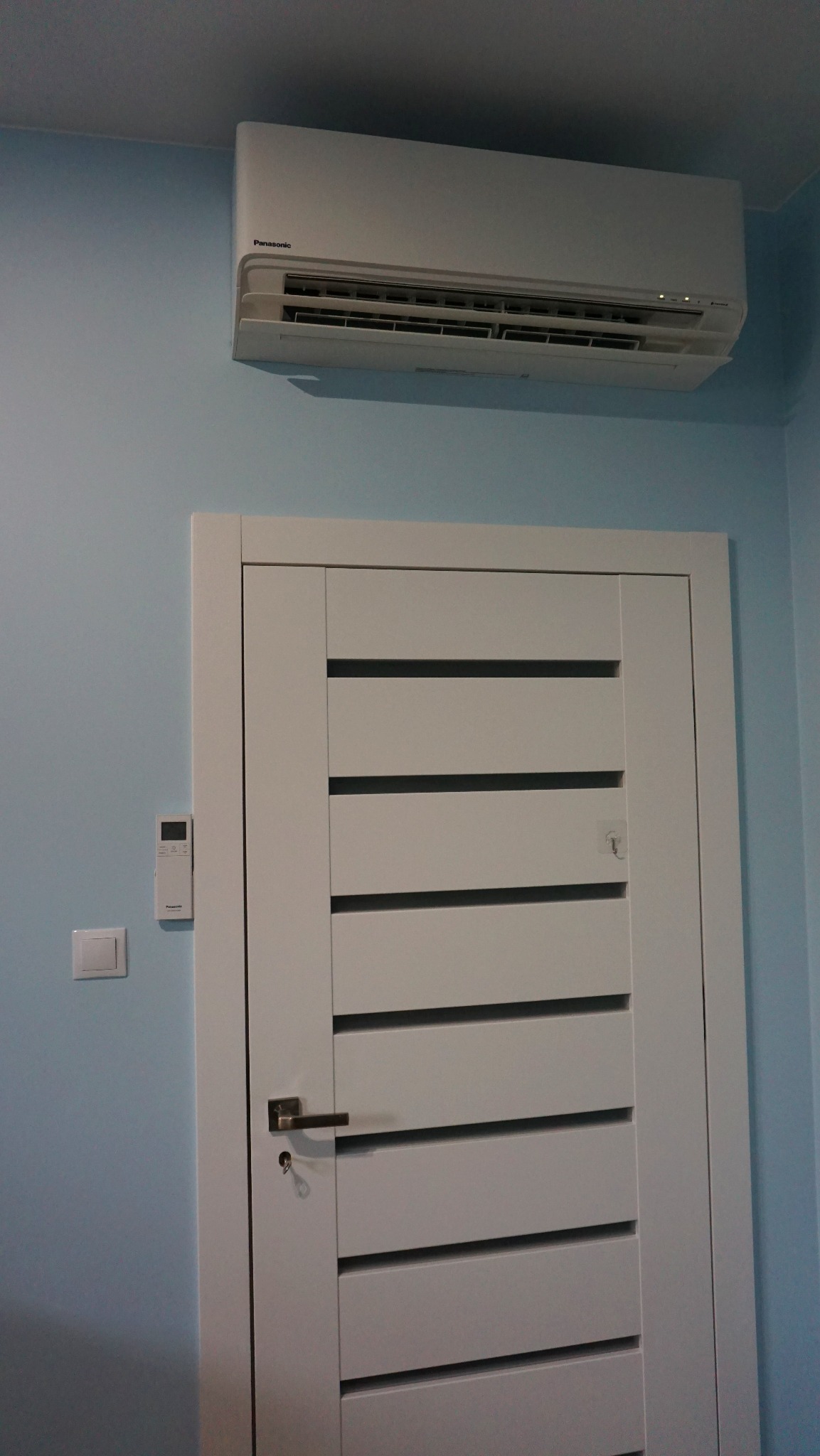 Biały klimatyzator Panasonic zamontowany nad nowoczesnymi, białymi drzwiami z poziomymi panelami na błękitnej ścianie. Widoczny pilot i włącznik światła.