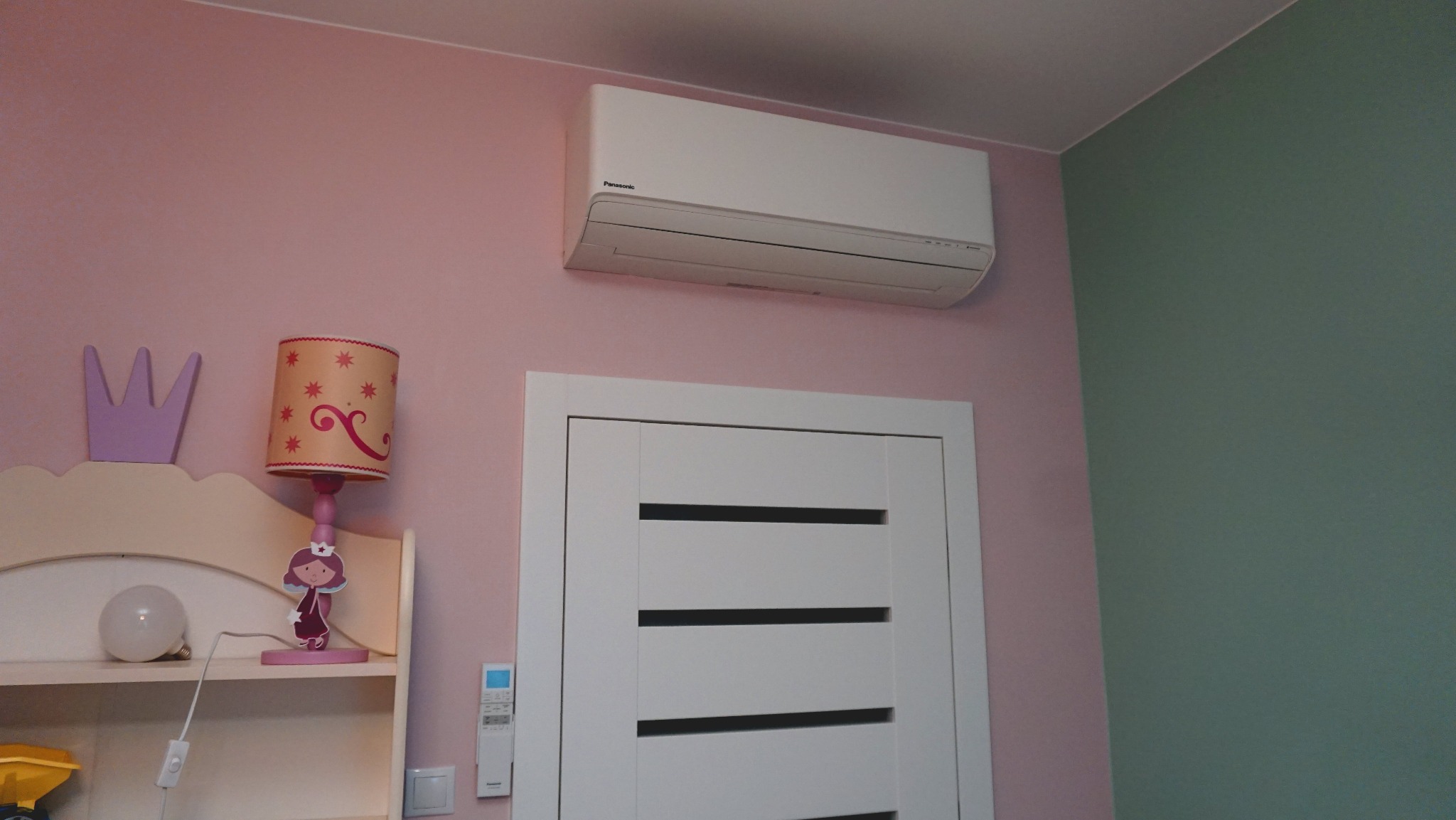 Biały klimatyzator Panasonic na różowej ścianie pokoju dziecięcego, nad białą ramą drzwi. Widoczny pilot i lampka nocna z motywem księżniczki.