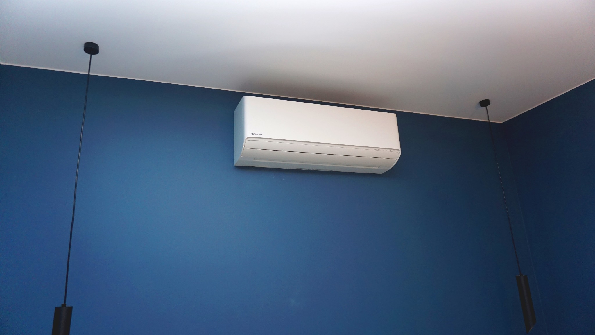 Biały klimatyzator Panasonic zamontowany na niebieskiej ścianie w pokoju z nowoczesnym oświetleniem. Widok z dołu, podkreślający estetykę instalacji.