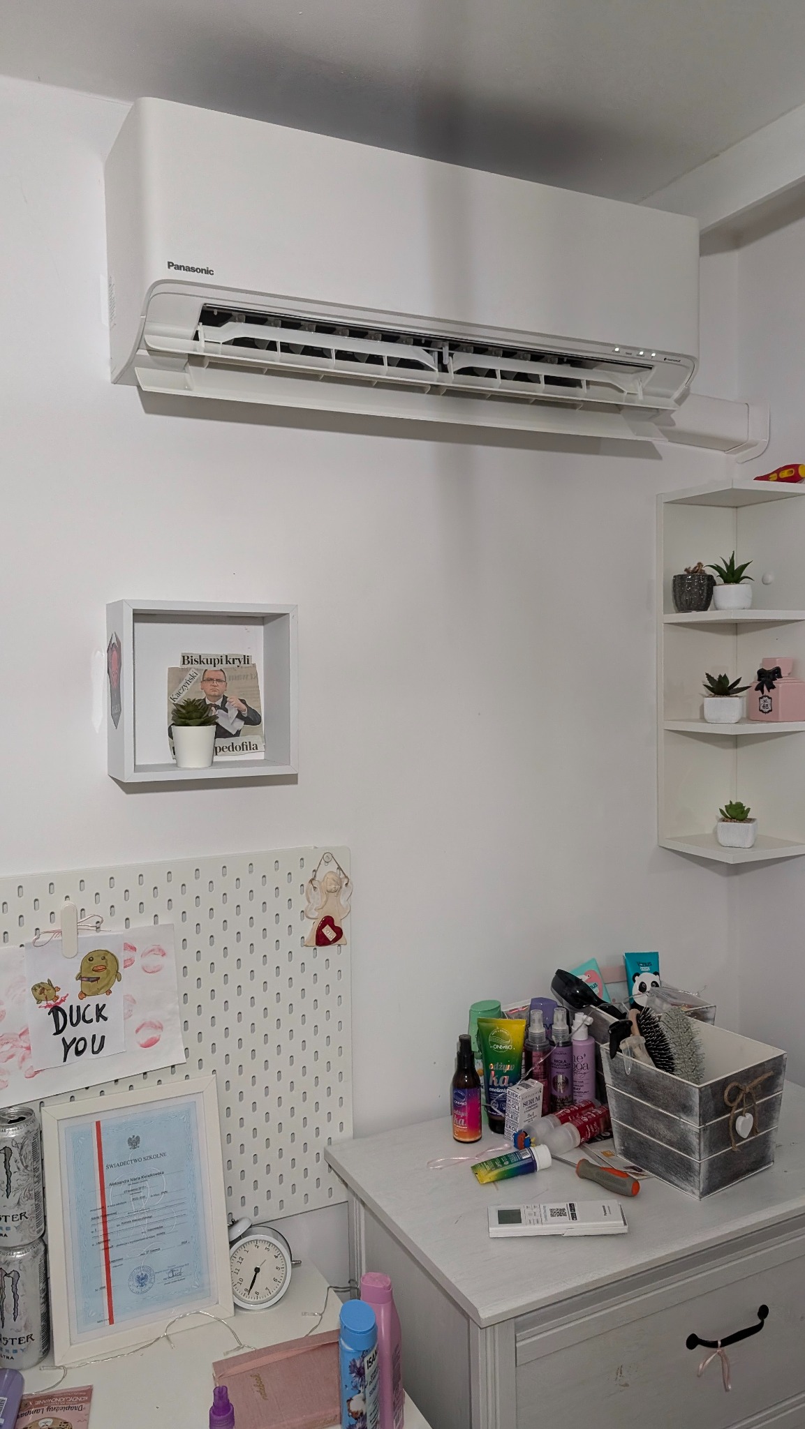 Biała klimatyzacja Panasonic zamontowana na ścianie w pokoju. Pod spodem biurko z różnymi przedmiotami i dekoracjami. Ujęcie z dołu, widoczne elementy wyposażenia wnętrza.