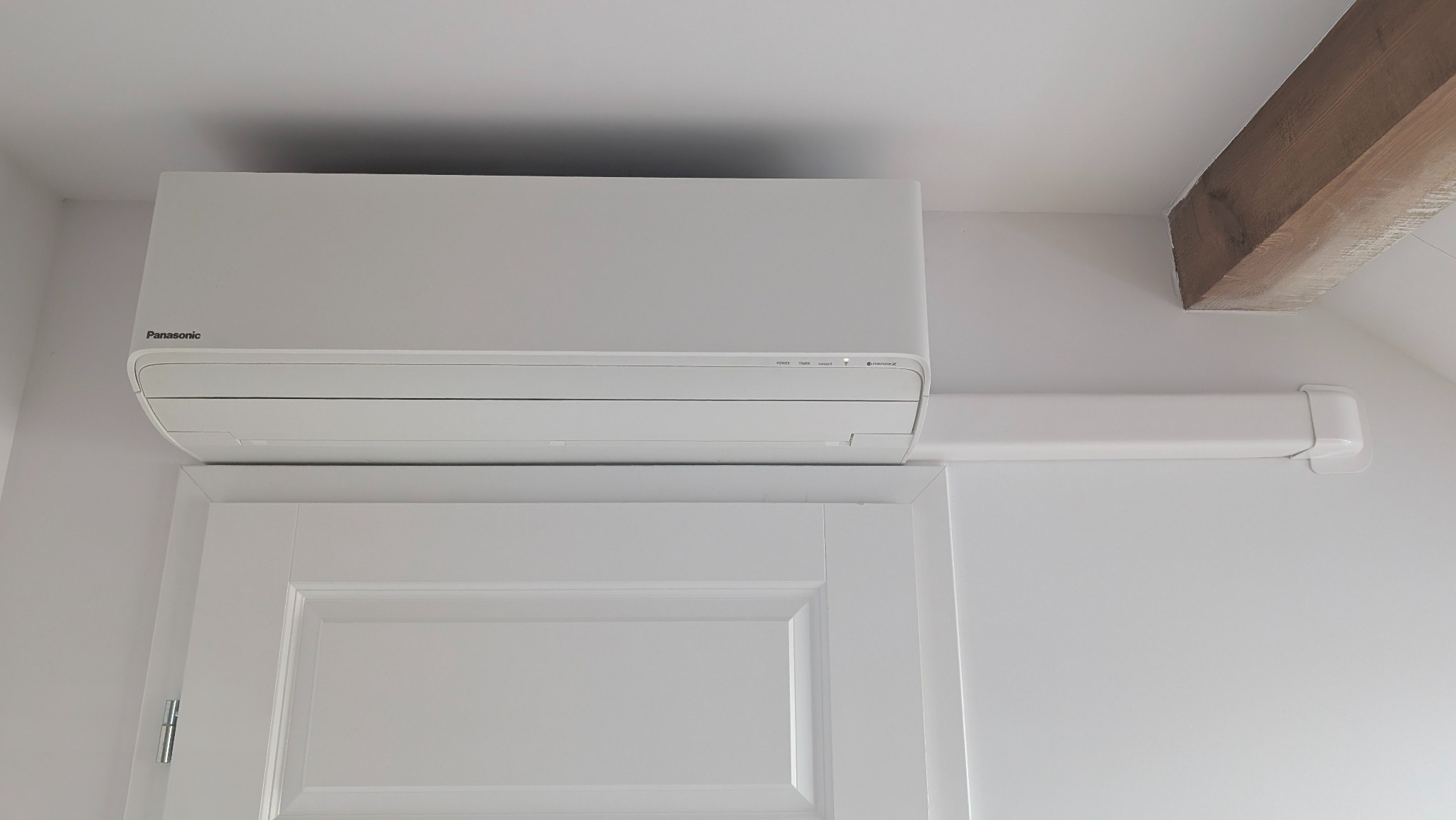 Biały klimatyzator Panasonic zamontowany nad drzwiami w pomieszczeniu z drewnianą belką stropową. Widoczne przewody w białej osłonie.